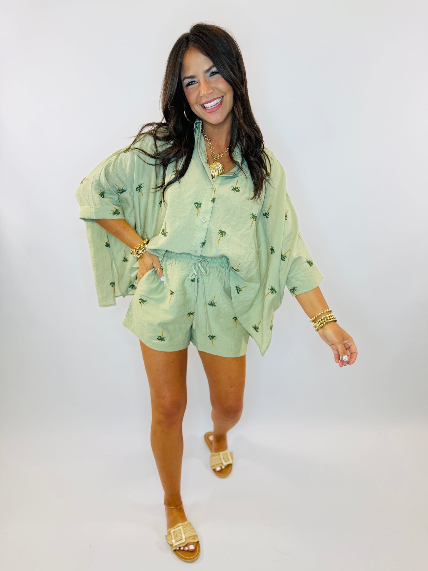 Woman wearing green palm tree print button-up top and matching shorts set with gold jewelry and sandals.