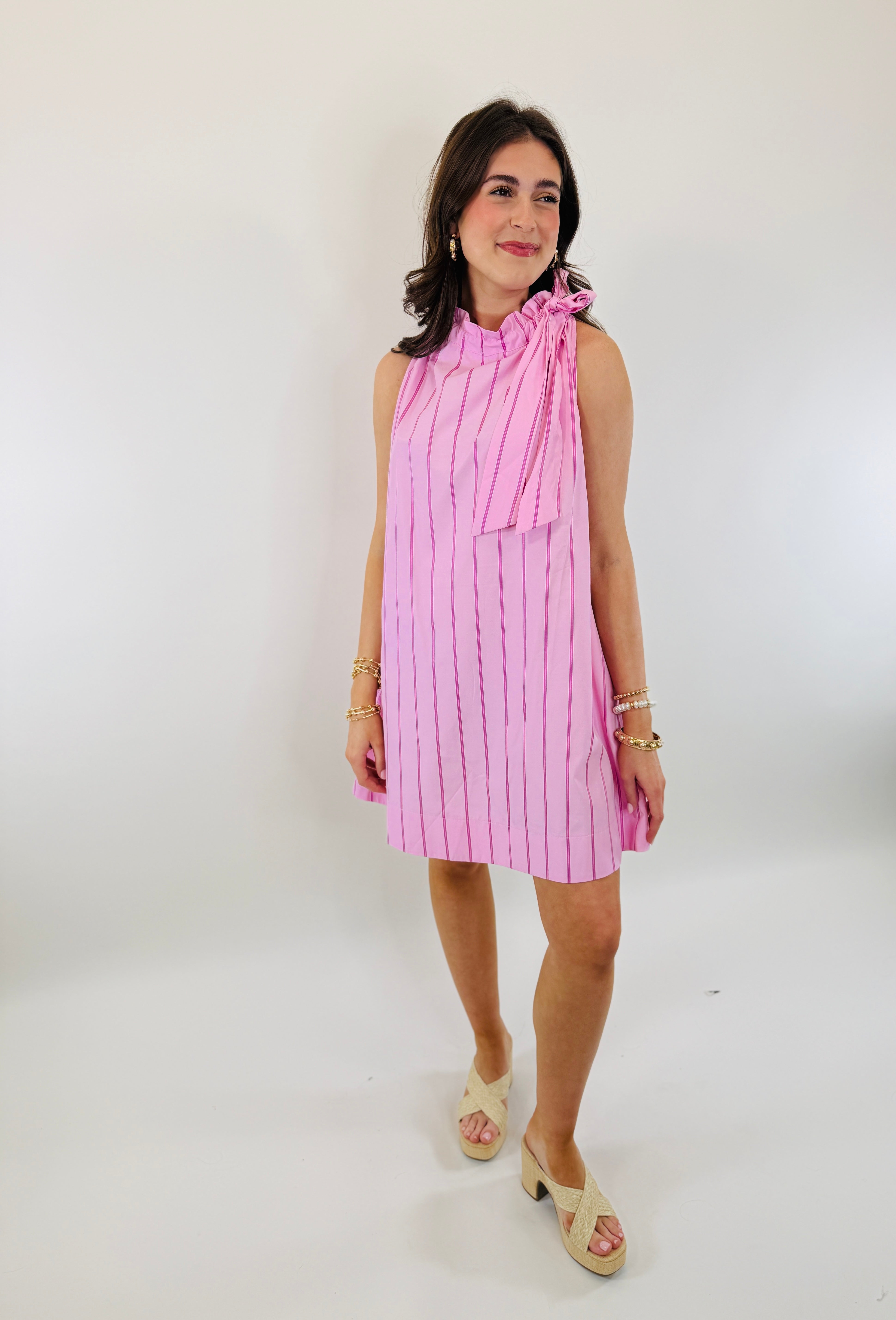 Woman wearing pink striped sleeveless Alice dress with tie neck detail and gold jewelry accessories.