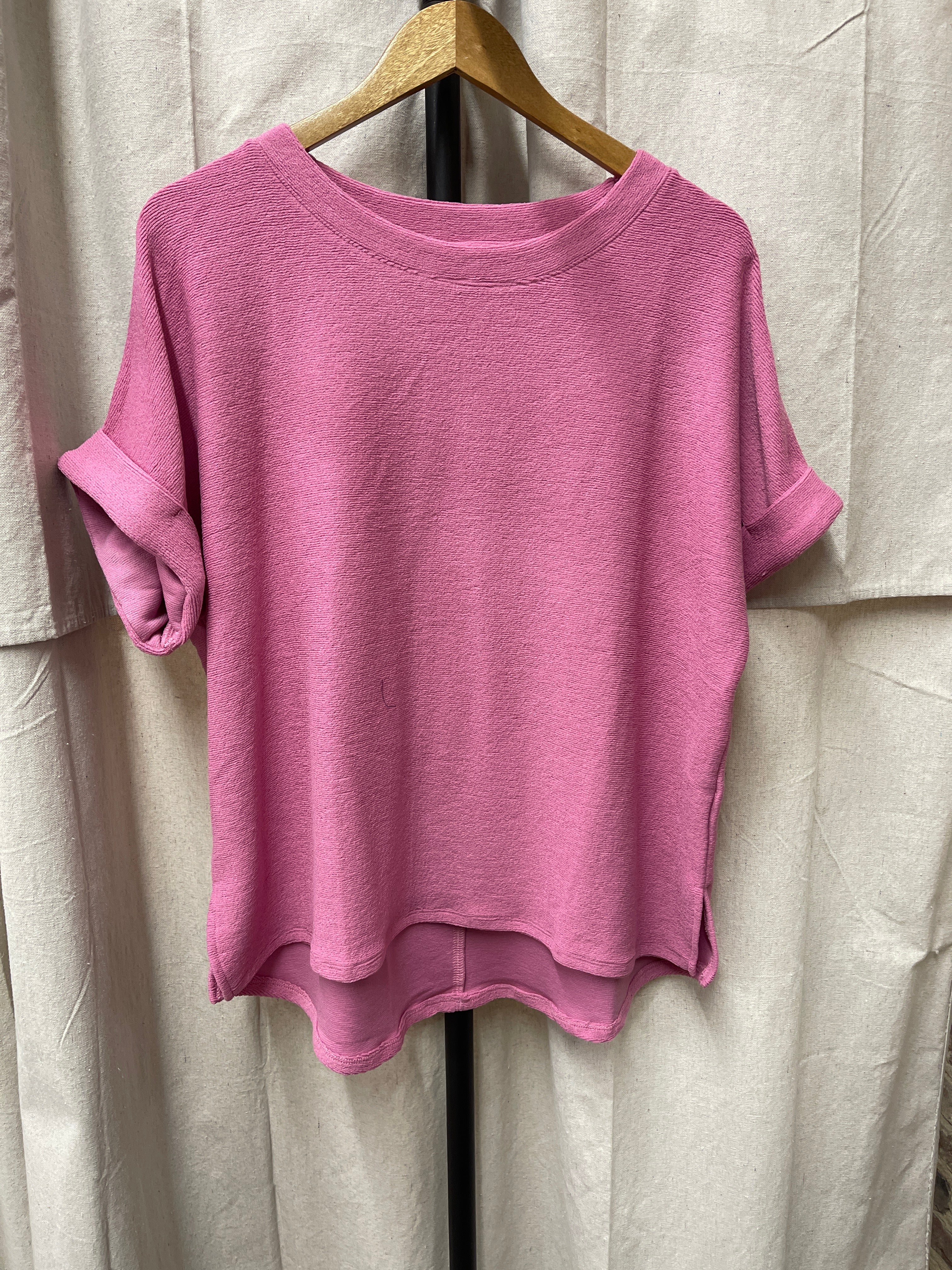 Pink relaxed-fit top with rolled sleeves and curved hem hanging on wooden hanger against beige backdrop.