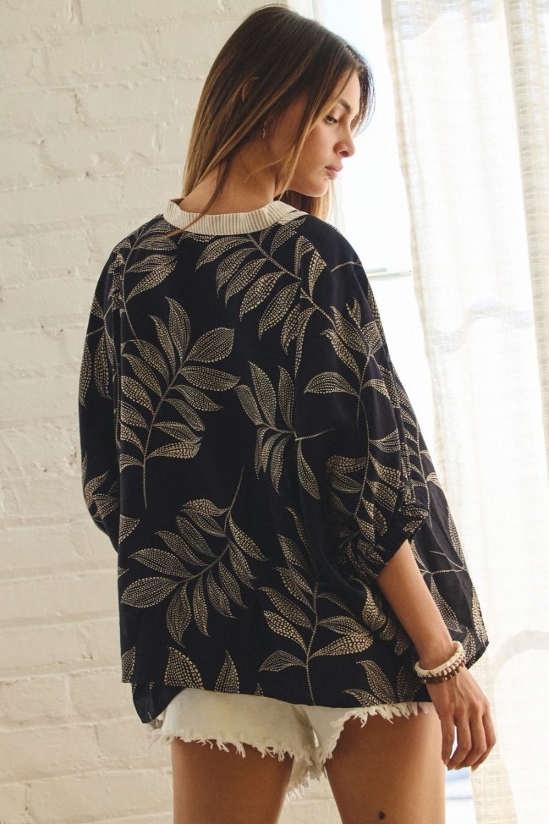 Woman wearing black oversized top with gold leaf pattern and cream ribbed neckline, paired with white denim shorts.