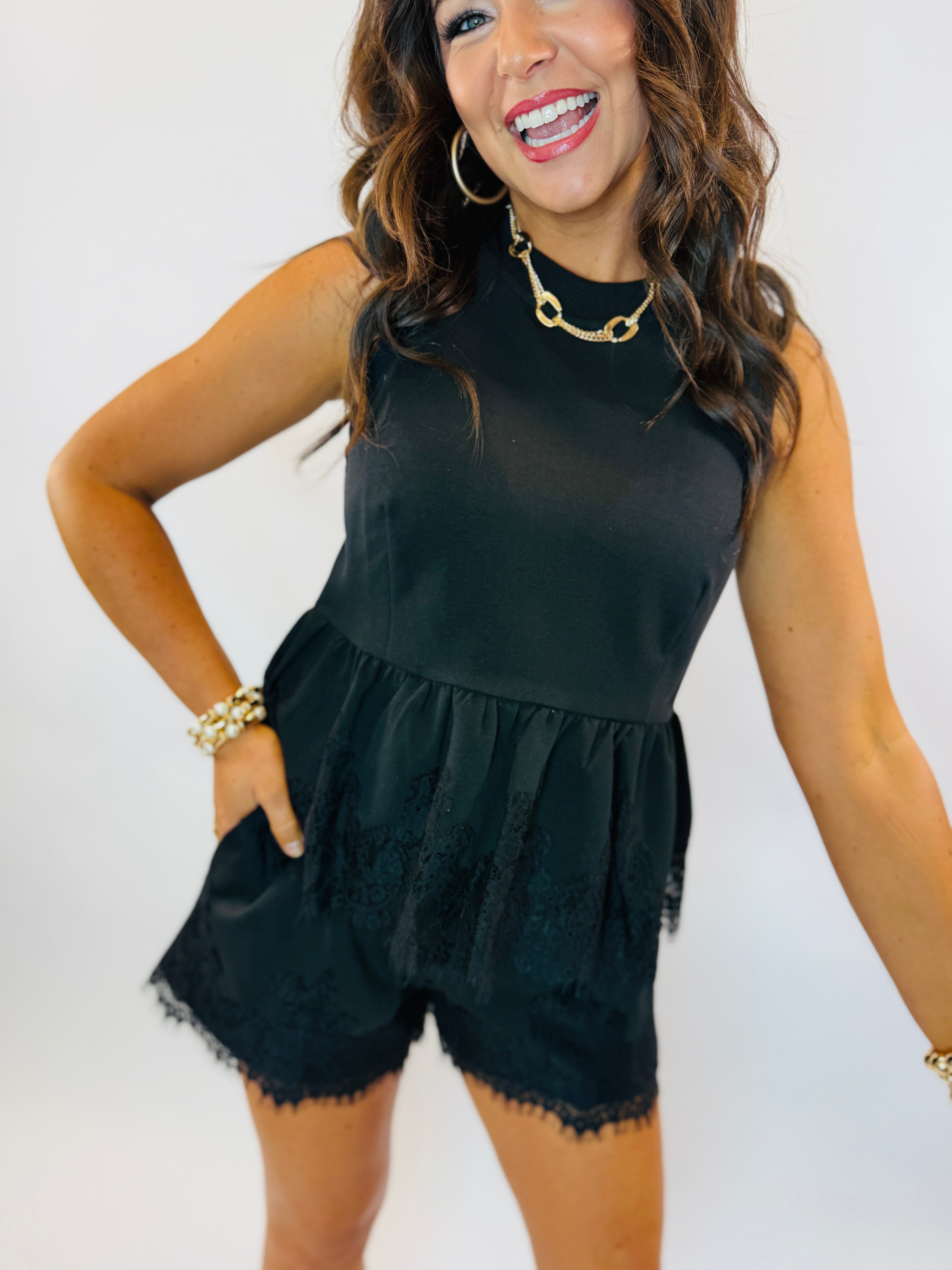 Woman wearing black sleeveless top with lace trim hem and gold chain necklace, smiling against white background.