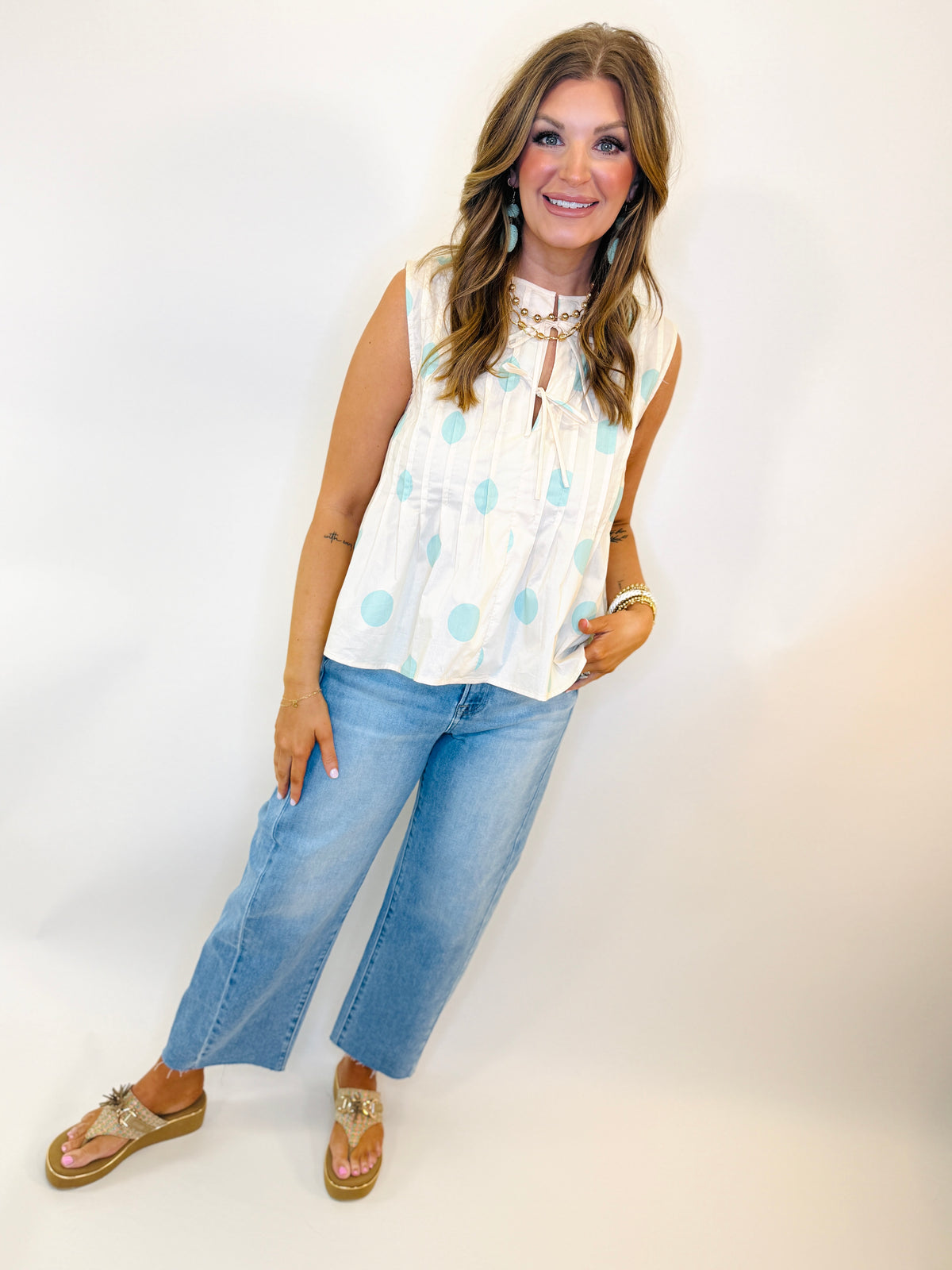 Woman wearing white sleeveless polka dot blouse with gold necklace, blue cropped jeans, and gold sandals.