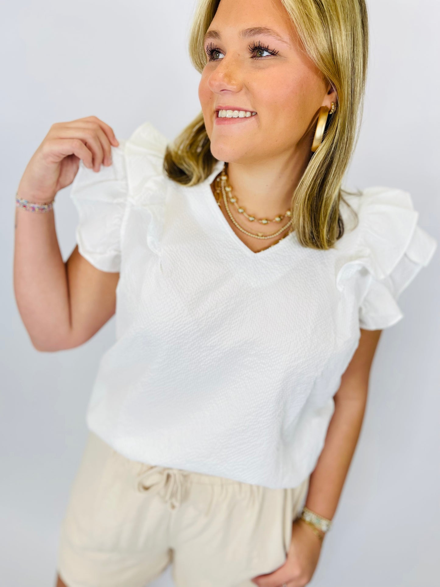 Woman wearing white textured Floriana top with ruffle sleeves and cream drawstring shorts, styled with layered necklaces.