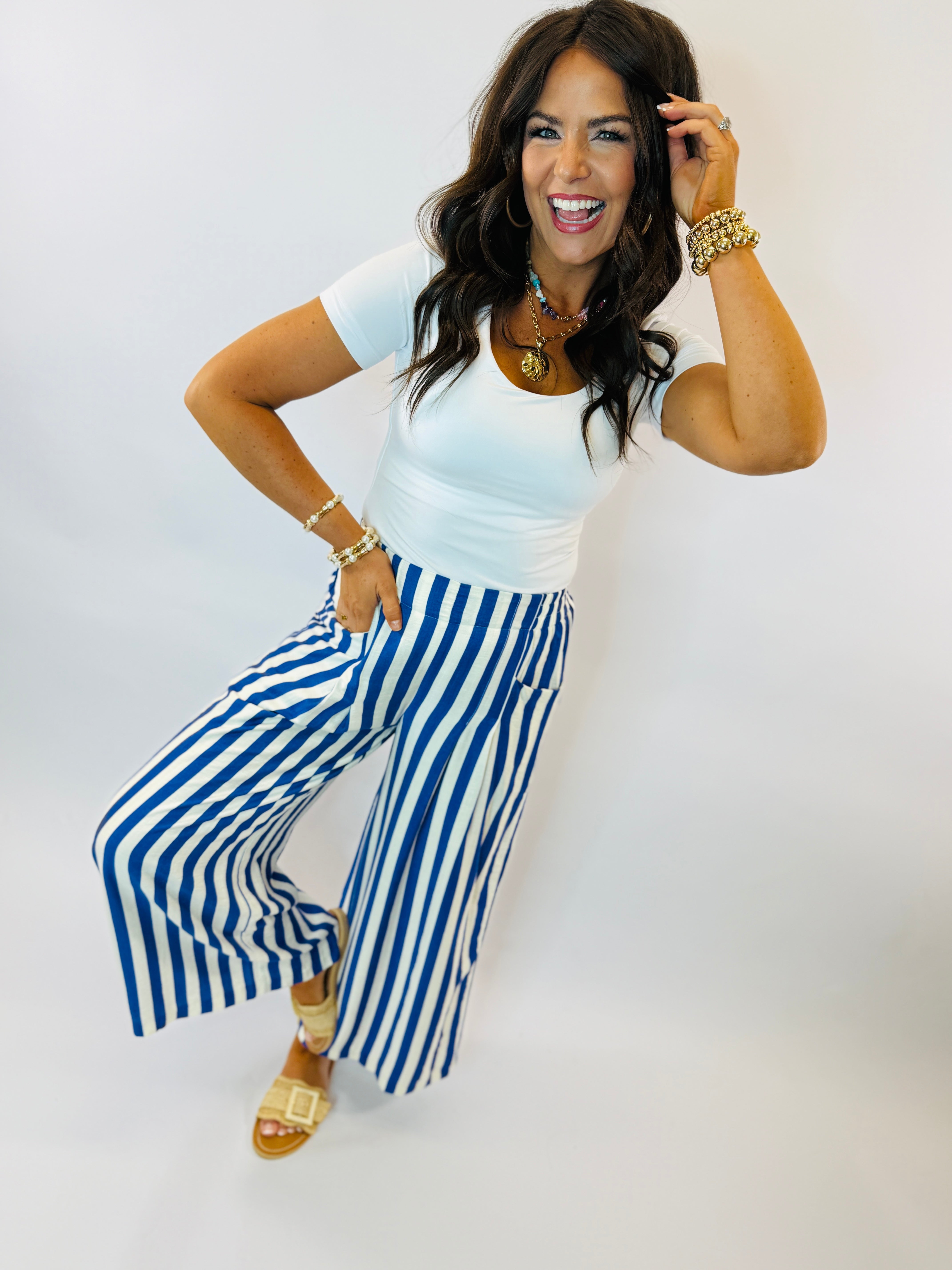 Woman wearing white top and blue and white striped wide-leg pants with gold accessories and sandals.