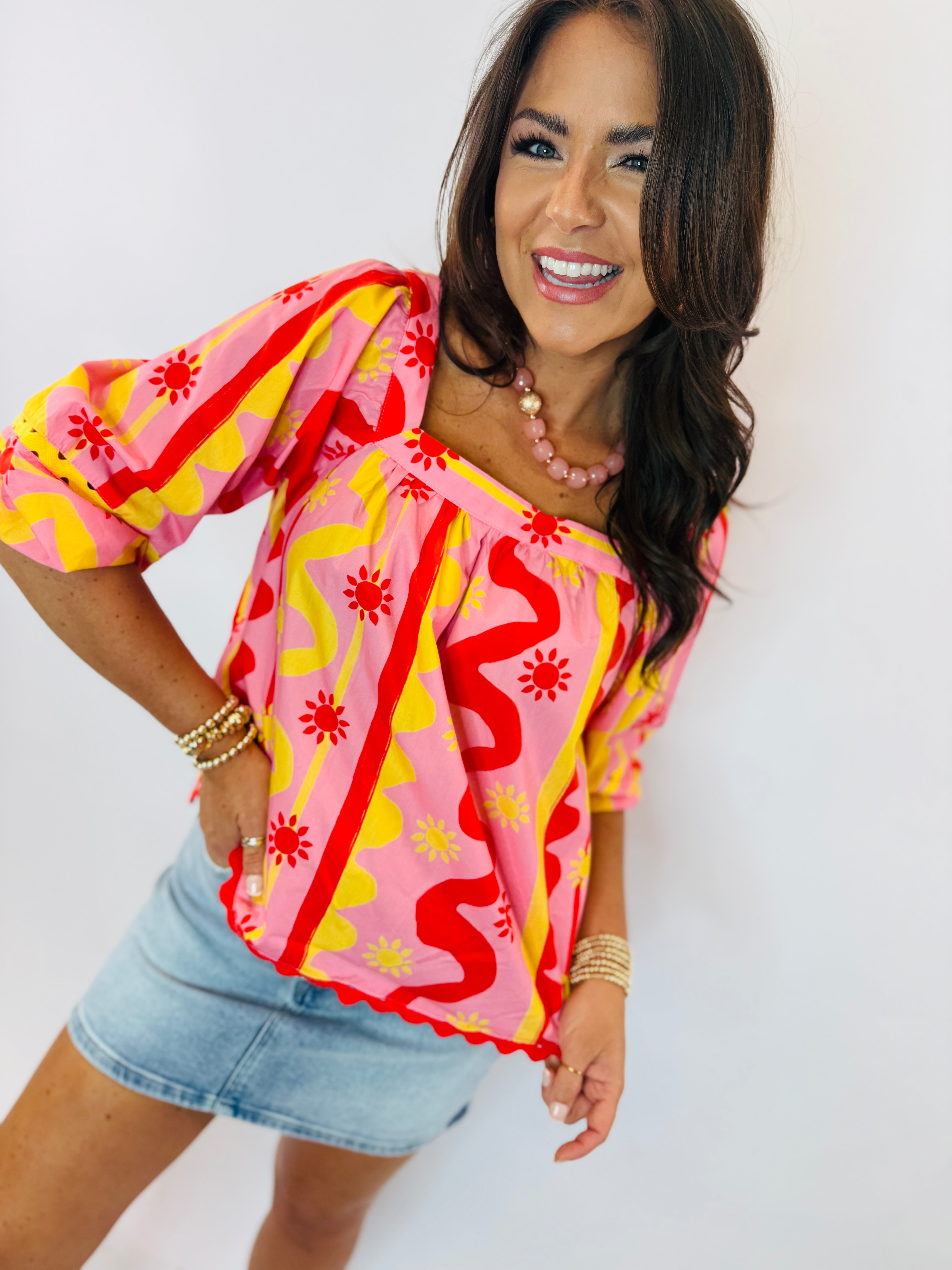 Woman wearing bright pink patterned Emmie top with puff sleeves and denim skirt, smiling at camera.