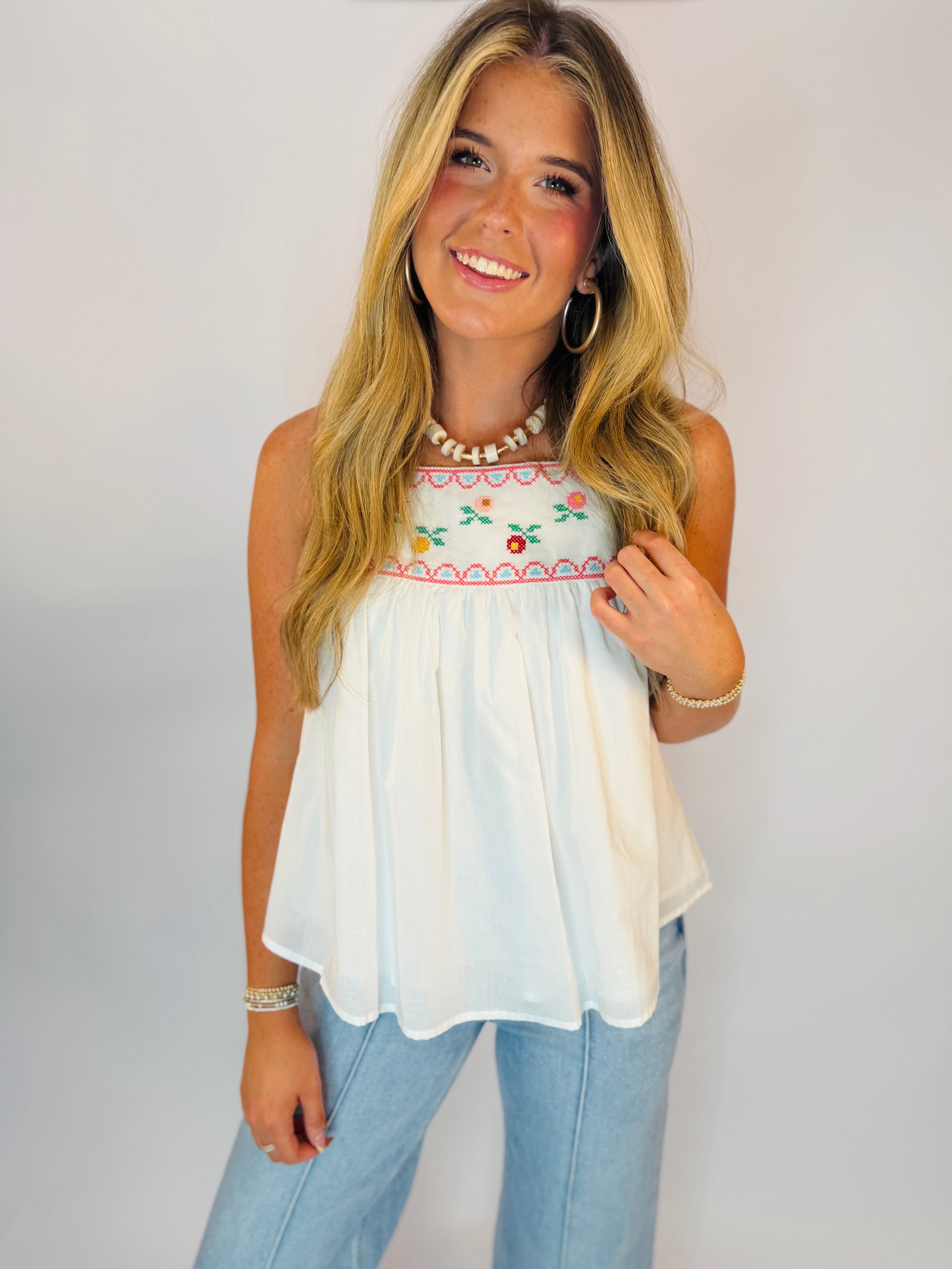 White sleeveless baby doll tank top with colorful cross-stitch floral embroidery at neckline, paired with light wash jeans.