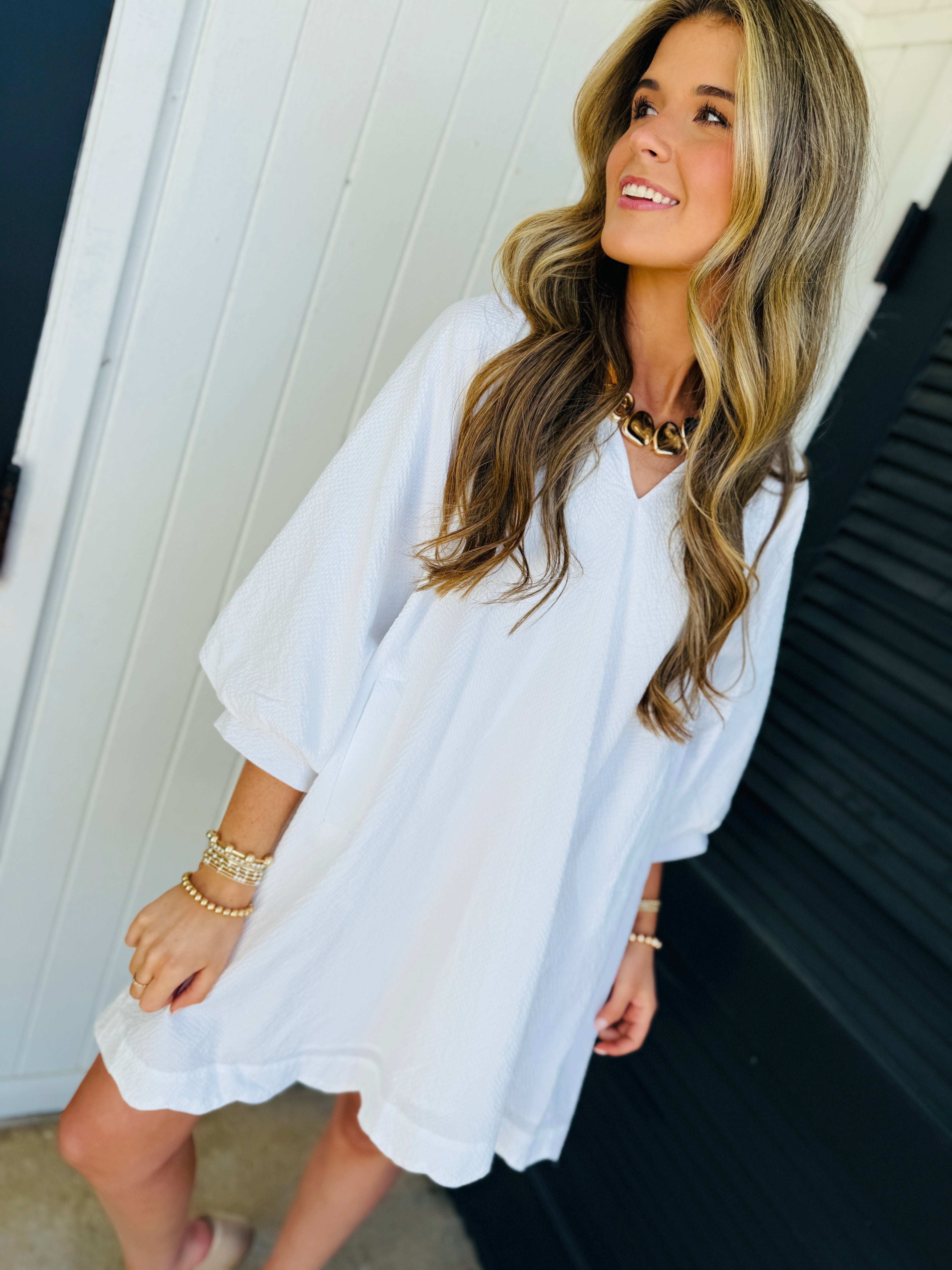 Woman wearing white casual v-neck dress with three-quarter sleeves, styled with gold jewelry and bracelets.