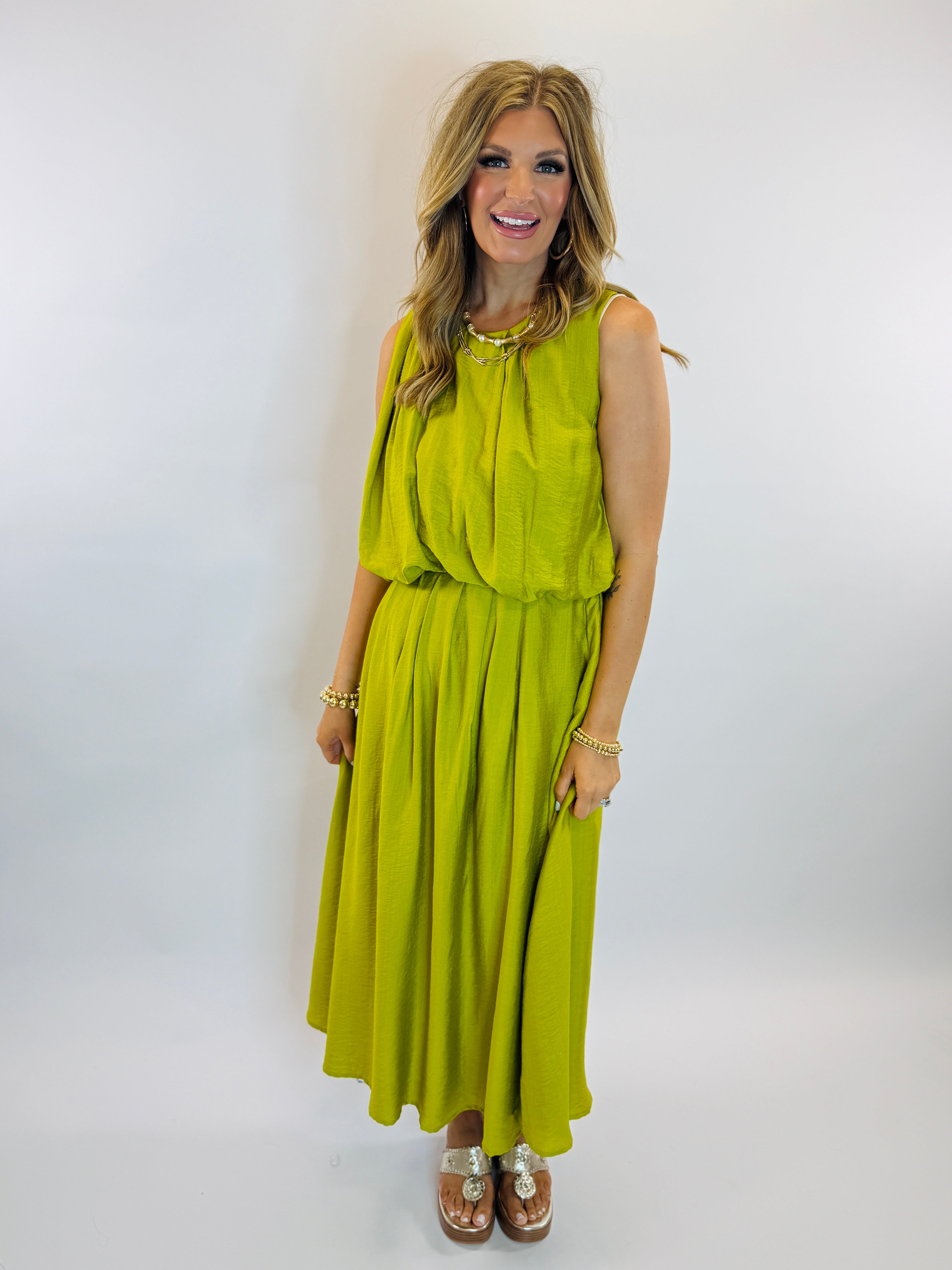 Woman wearing bright green sleeveless midi dress with gathered waist and flowing skirt, styled with gold jewelry and sandals.