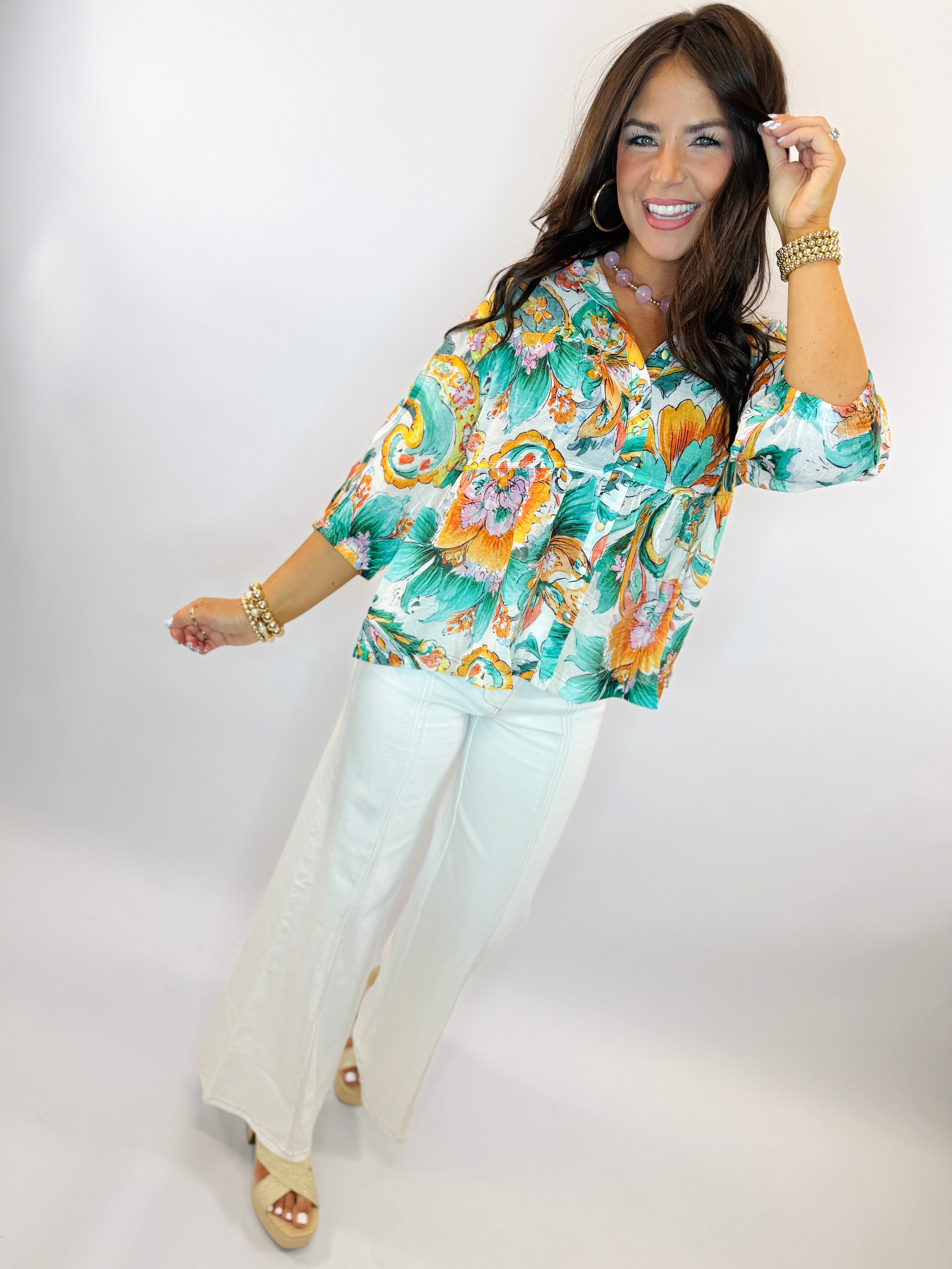 Woman wearing colorful floral print blouse with white pants and gold jewelry, smiling and posing