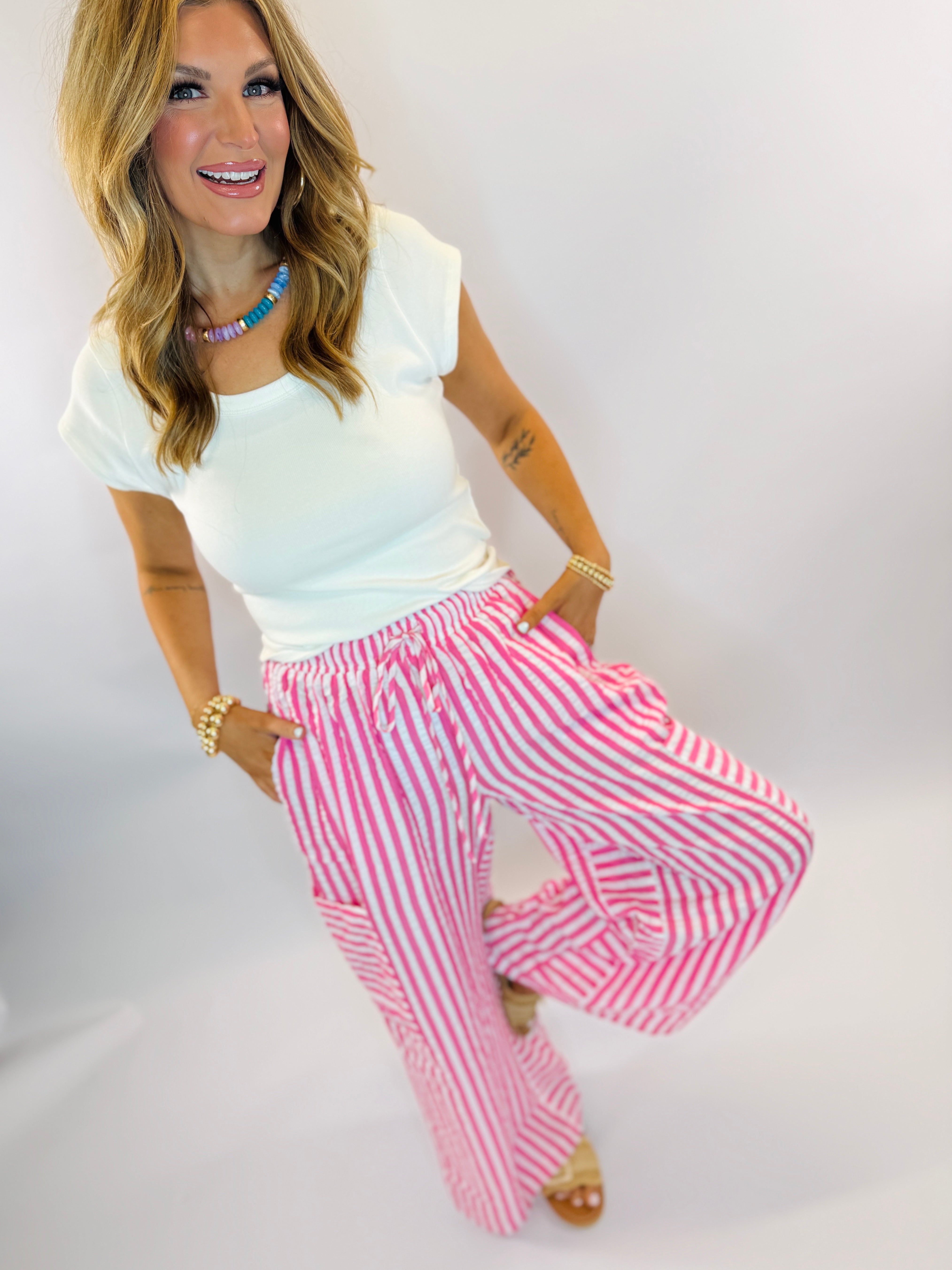 Woman wearing white top and pink striped wide-leg palazzo pants with elastic waistband, styled with colorful jewelry.