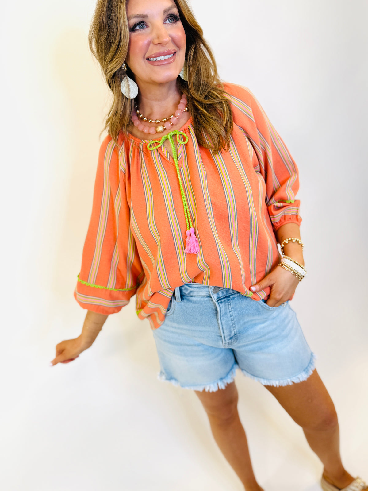 Woman wearing orange striped peasant blouse with green tassel ties and denim shorts, styled with layered jewelry.