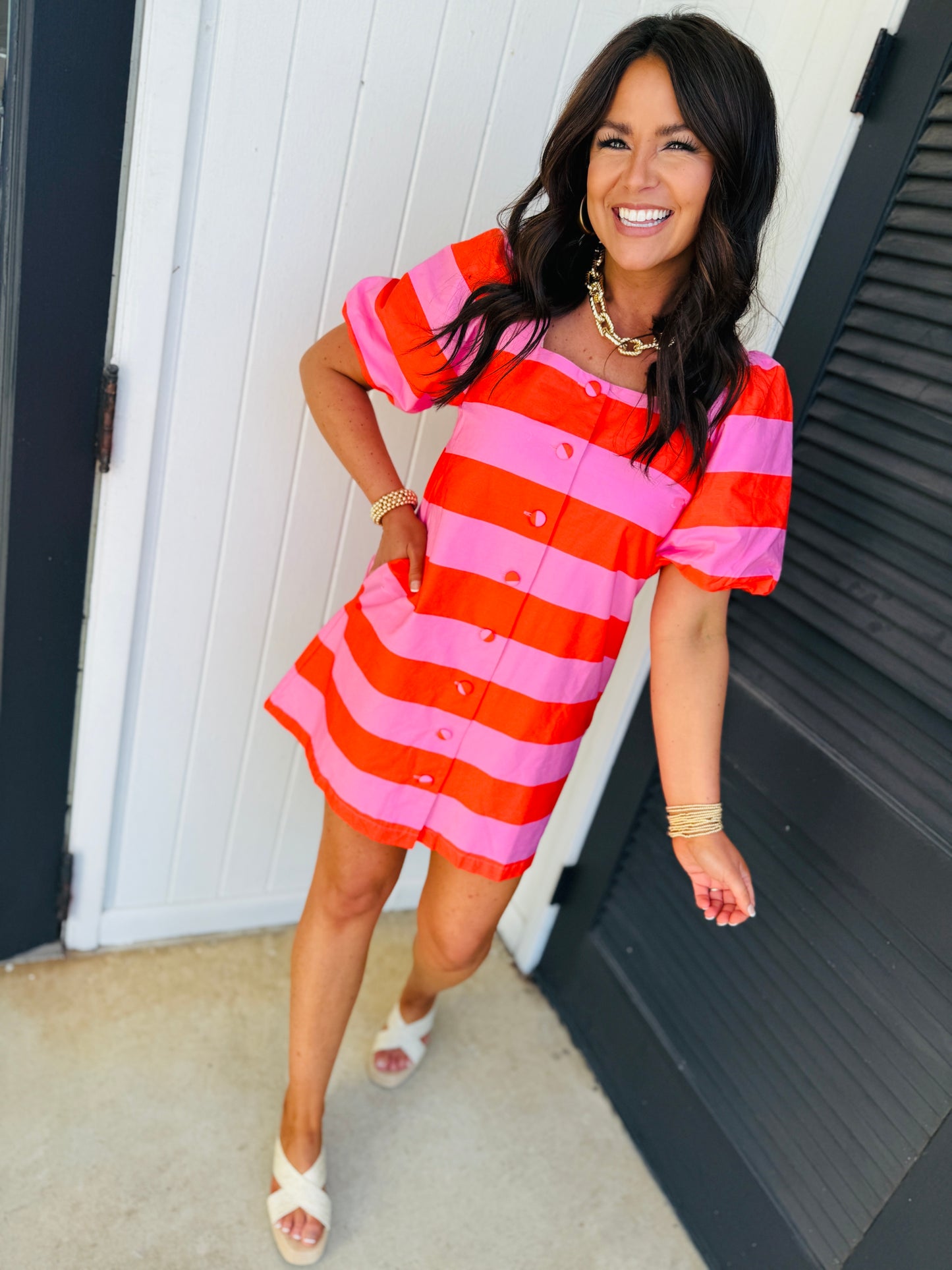 Woman wearing red and pink striped button-front dress with short sleeves, paired with white sandals and gold jewelry.