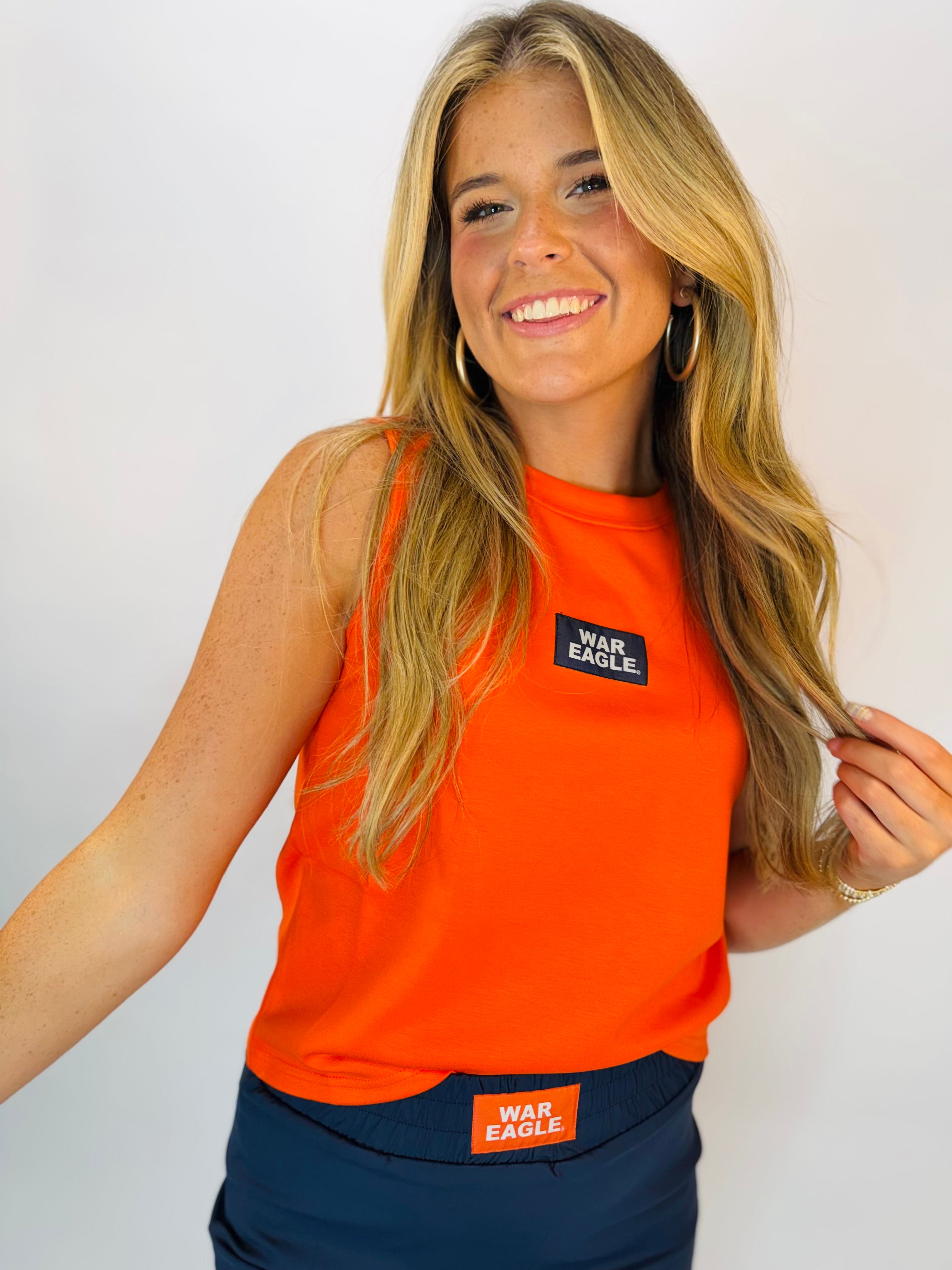 Woman wearing orange sleeveless tank top with navy "War Eagle" patch logo on chest.