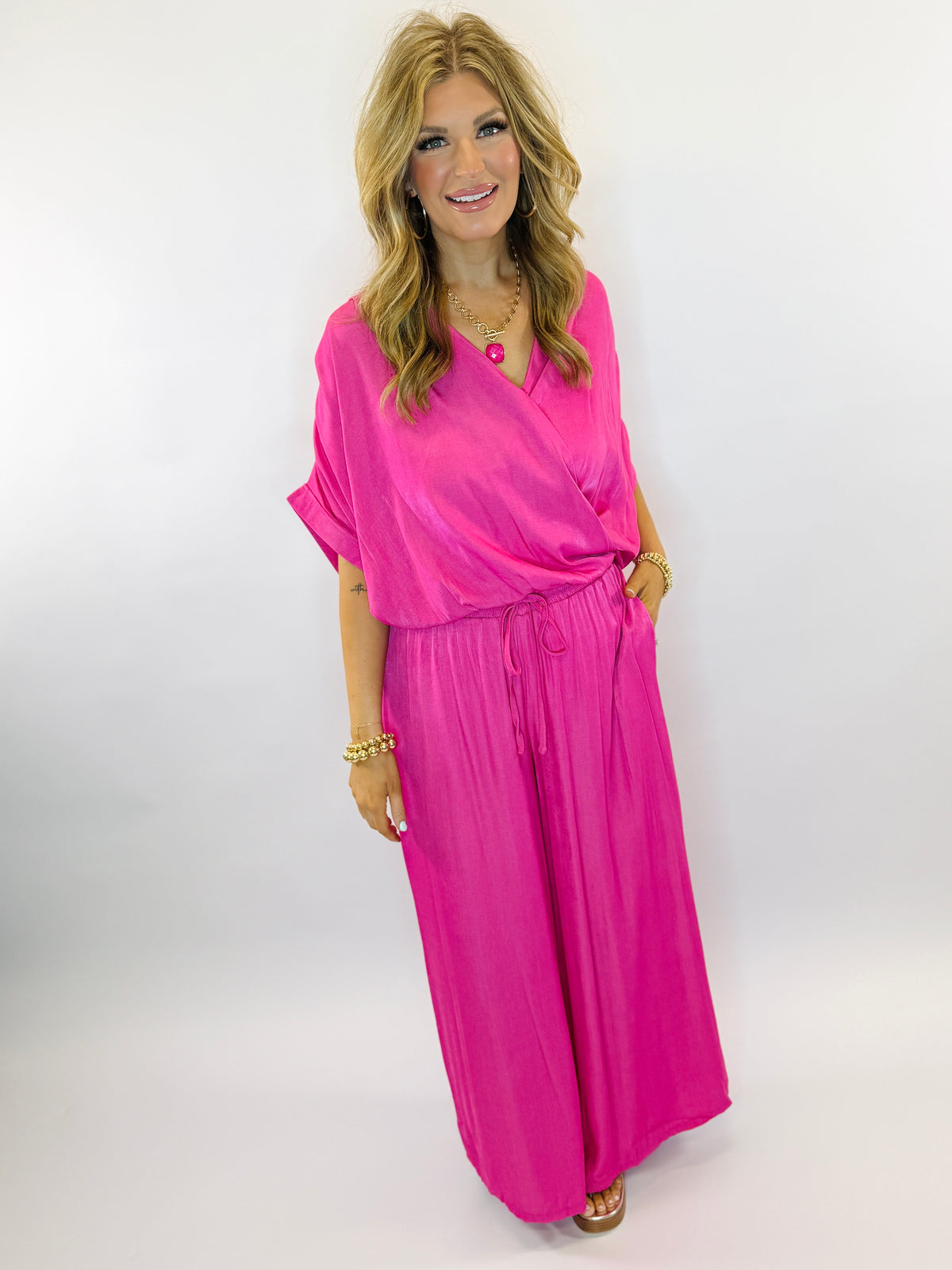 Woman wearing bright pink wrap-style top with matching wide-leg pants, styled with gold jewelry and blonde wavy hair.