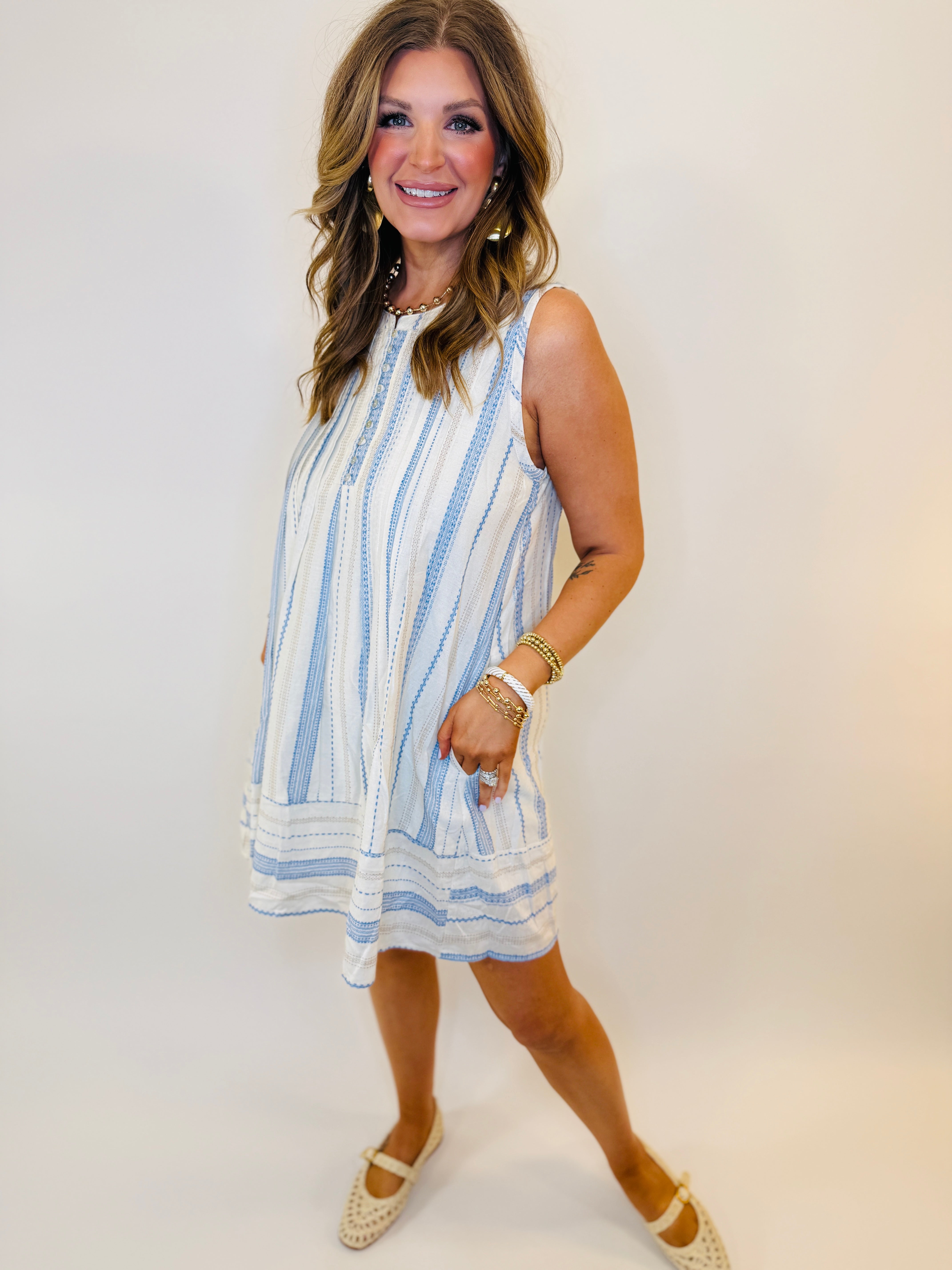 Woman wearing white sleeveless dress with blue vertical stripes and tiered hem, styled with gold jewelry and cream flats.