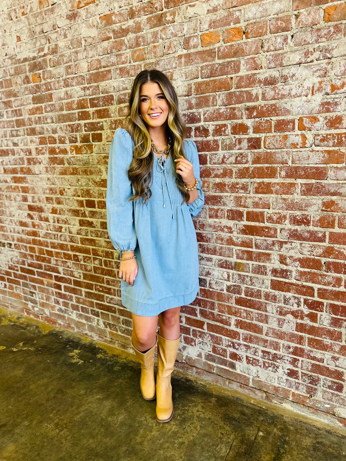 Woman wearing light blue long-sleeved dress with tie neckline and tan boots against brick wall.