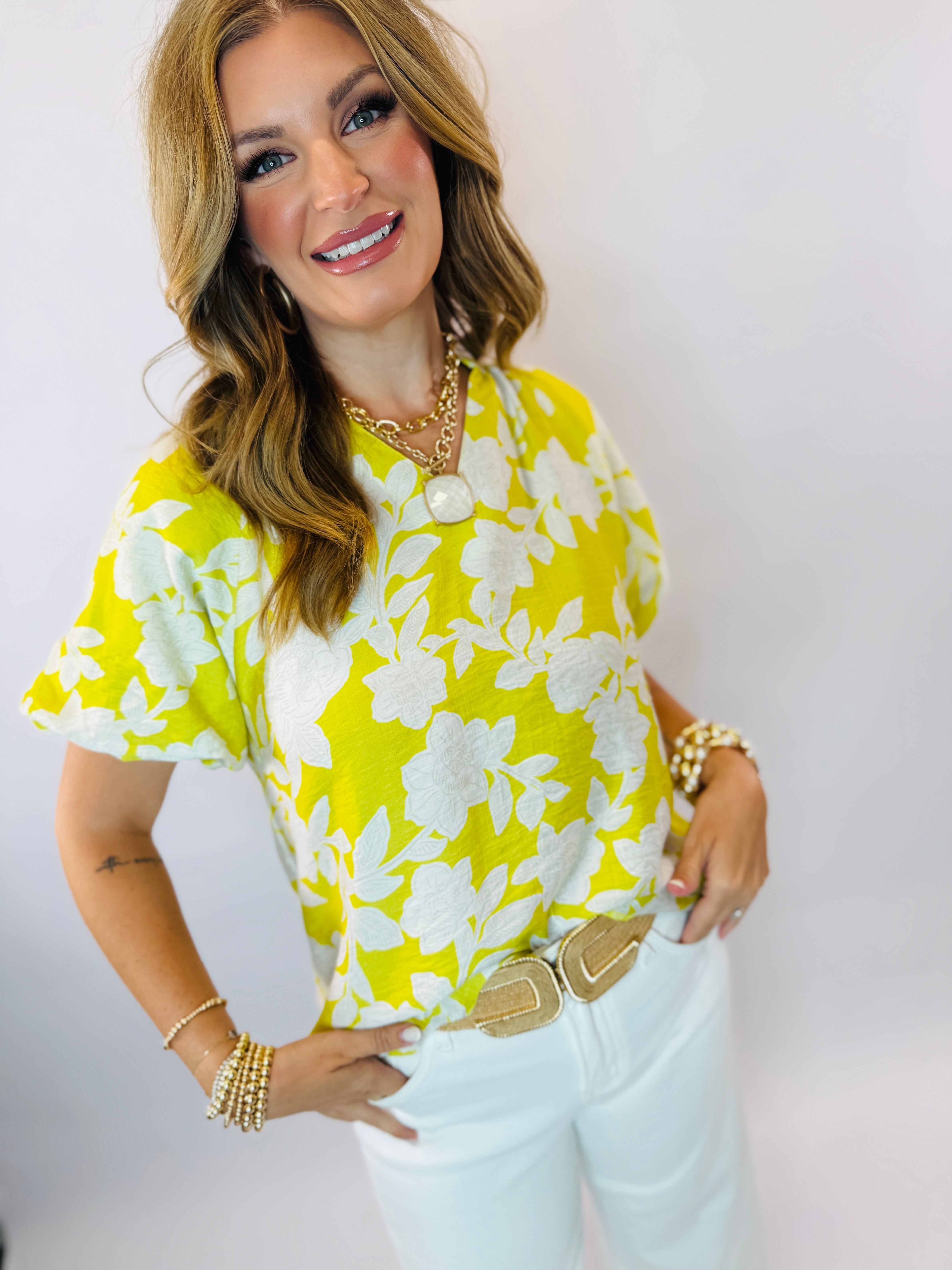Woman wearing yellow and white floral print Tessie top with gold jewelry and white pants