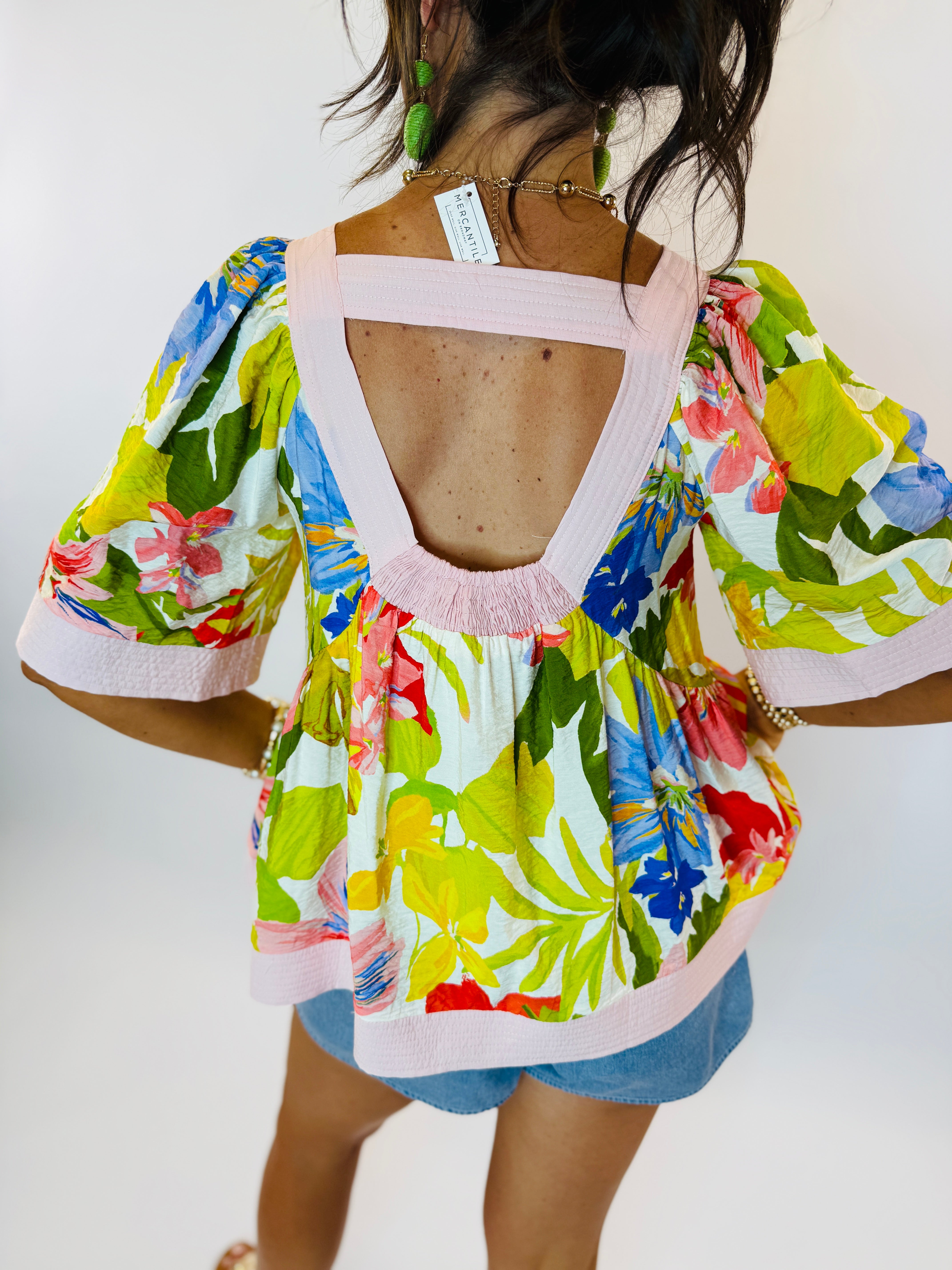 Colorful tropical floral print blouse with open back detail, pink trim, and flowing sleeves worn with denim shorts.