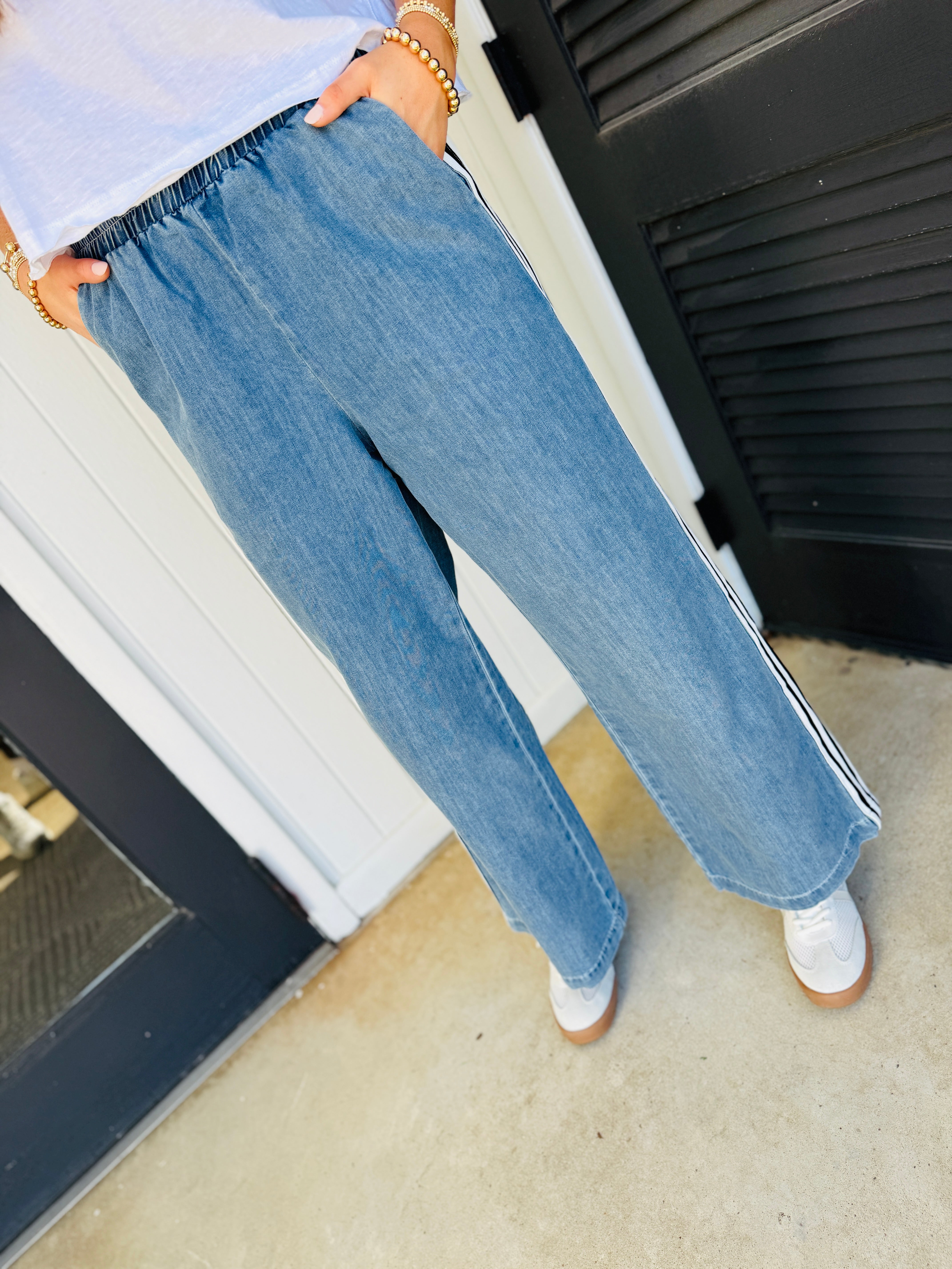 Woman wearing light blue relaxed-fit denim pants with elastic waistband and side stripe detail, paired with white sneakers.