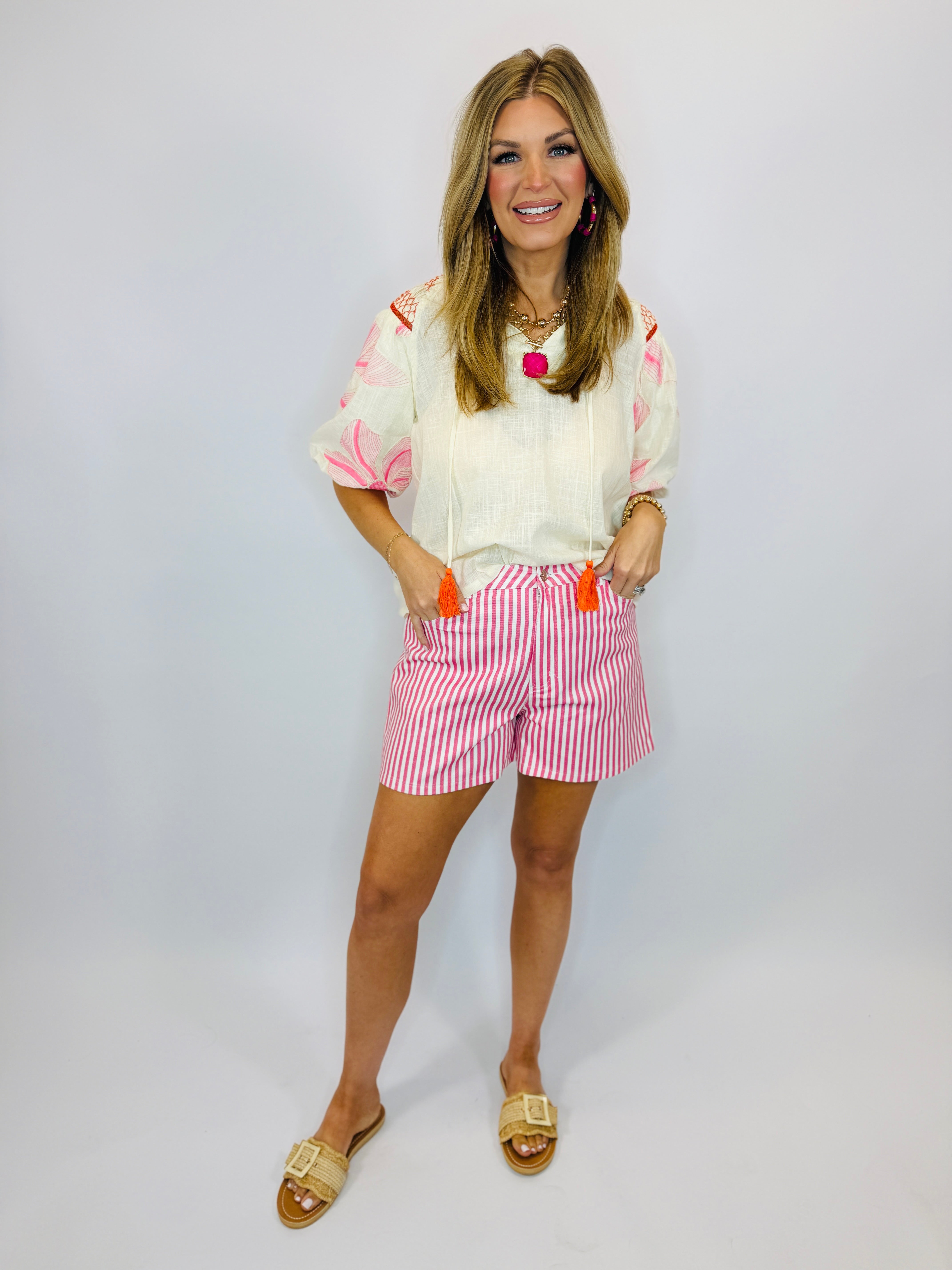 Woman wearing white embroidered Vera top with pink floral sleeves, pink striped shorts, and gold sandals