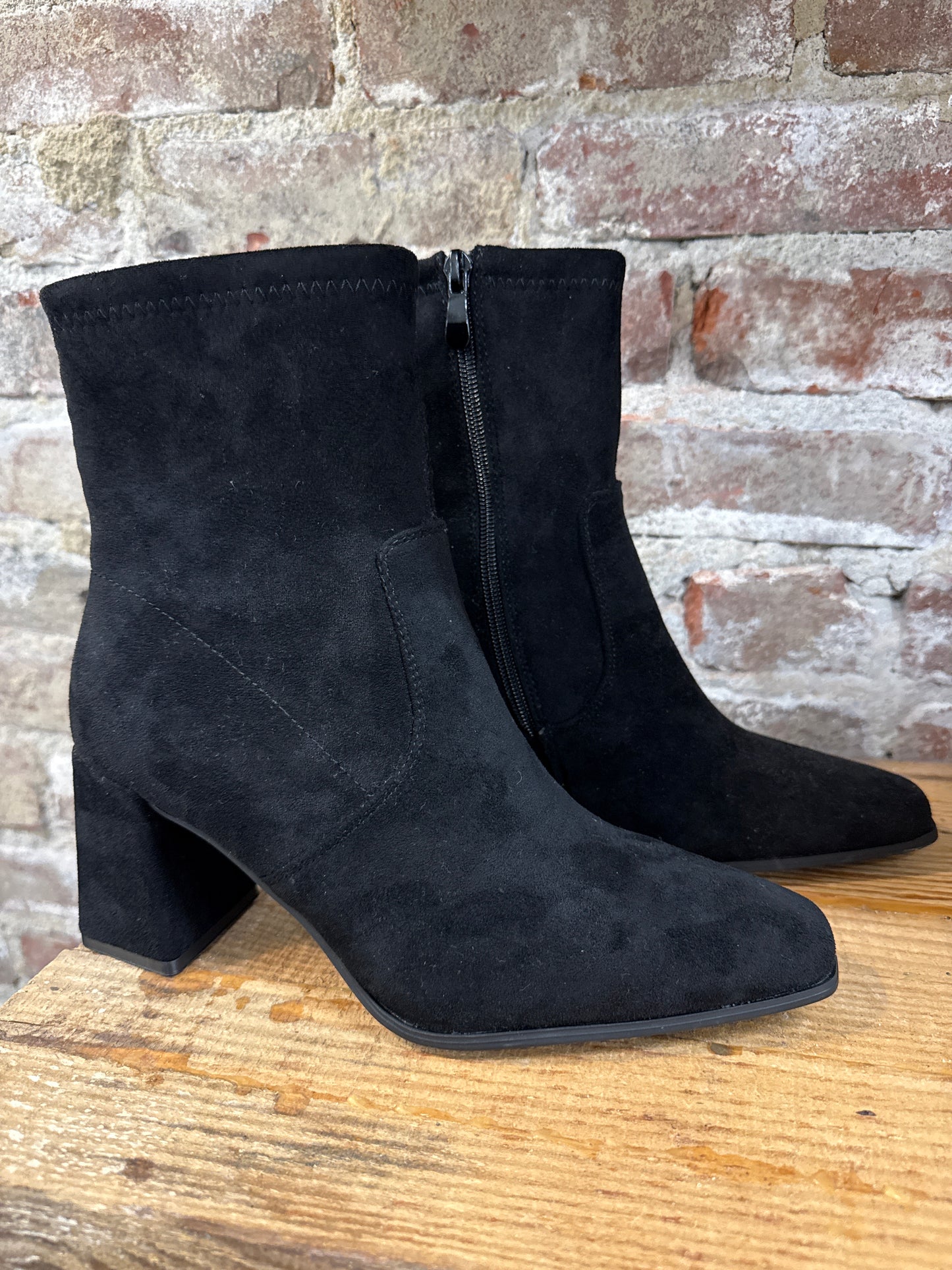 Black suede ankle boots with block heels and side zippers displayed on wooden surface against brick wall