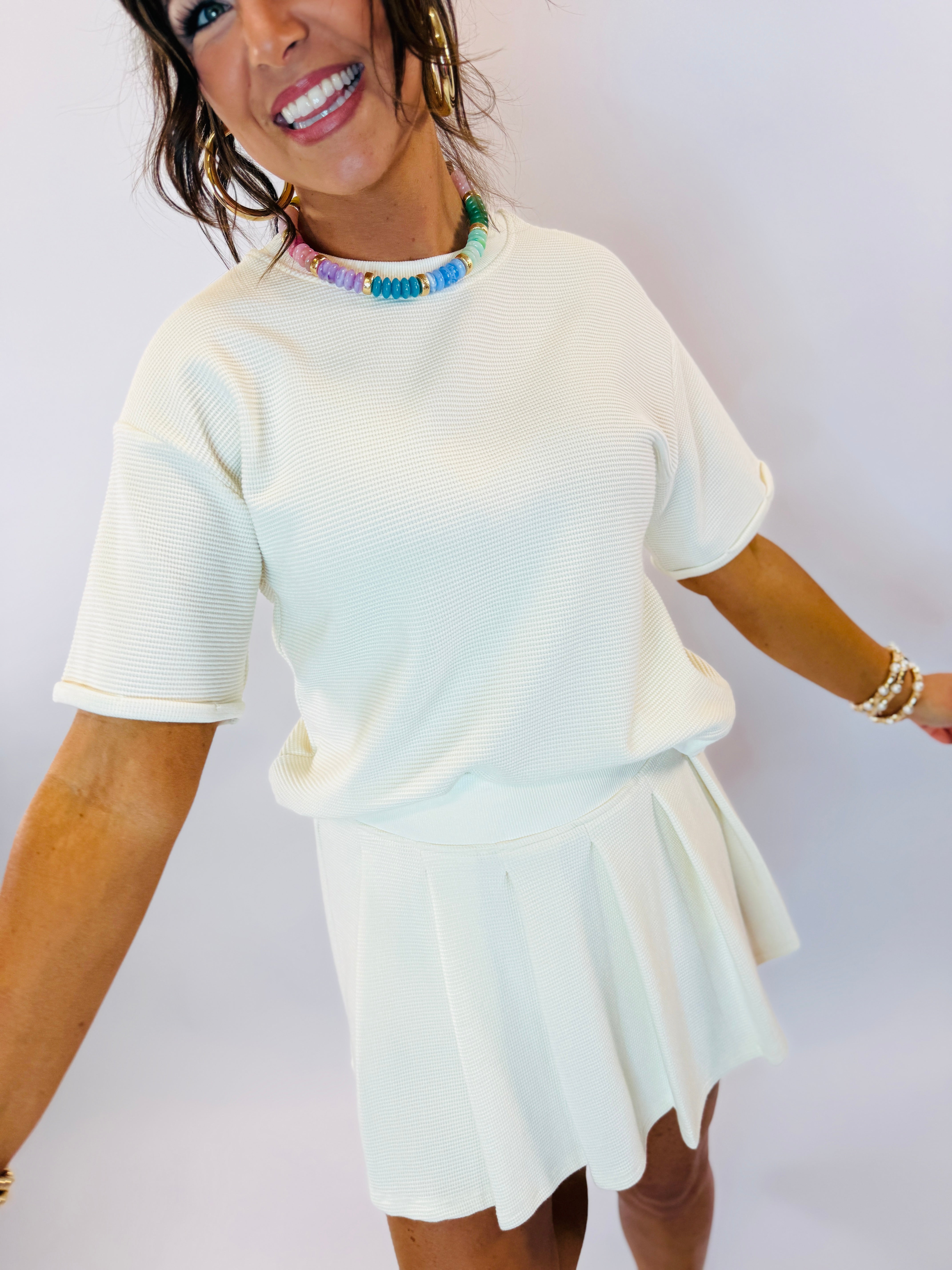 Woman wearing white textured matching top and skort set with colorful beaded necklace and bracelets.