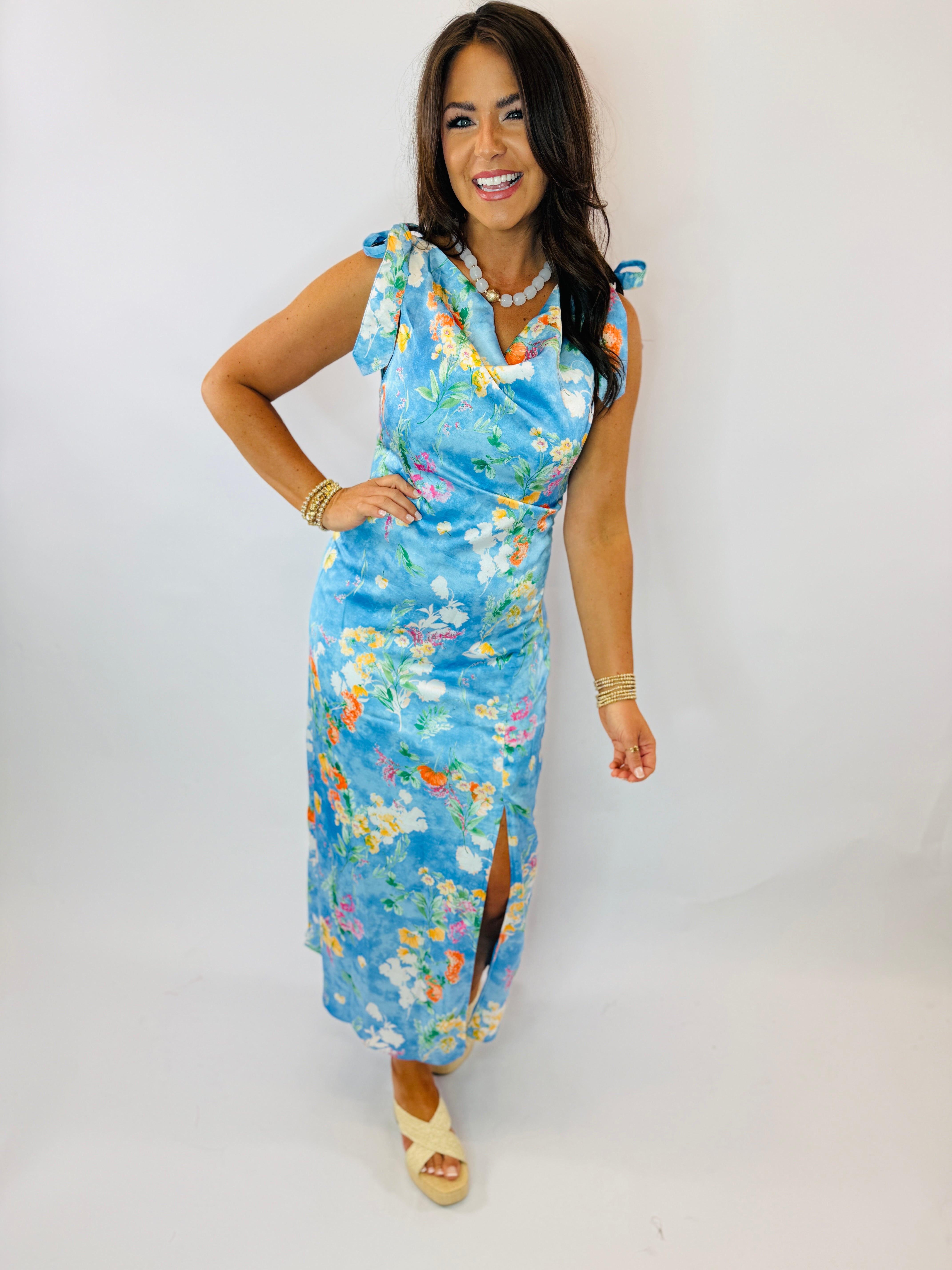 Woman wearing blue floral Ava dress with tie shoulders, pearl necklace, and platform sandals against white background.