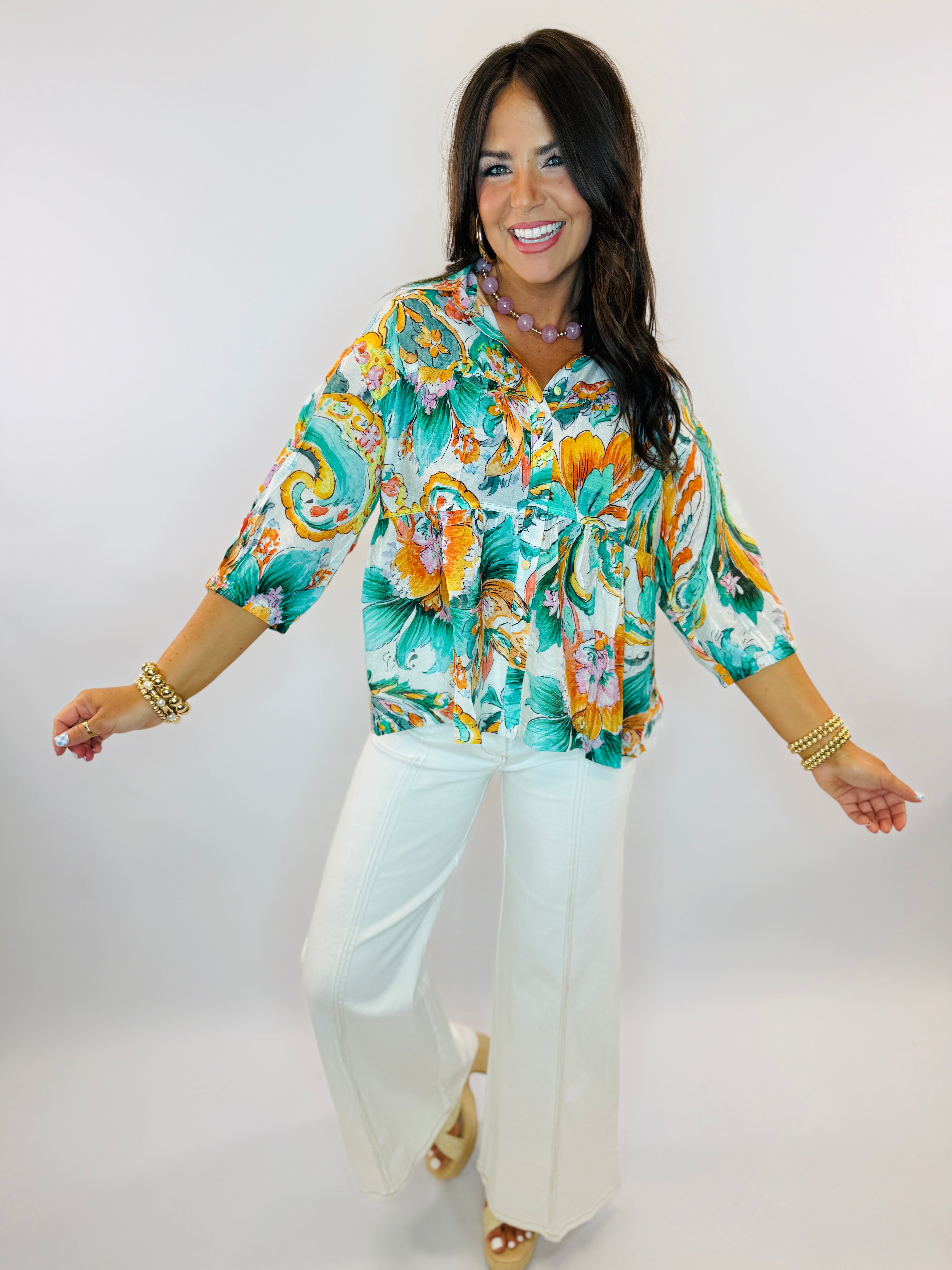 Woman wearing colorful tropical floral button-up shirt with white flare jeans, gold bracelets and necklace, smiling with a...