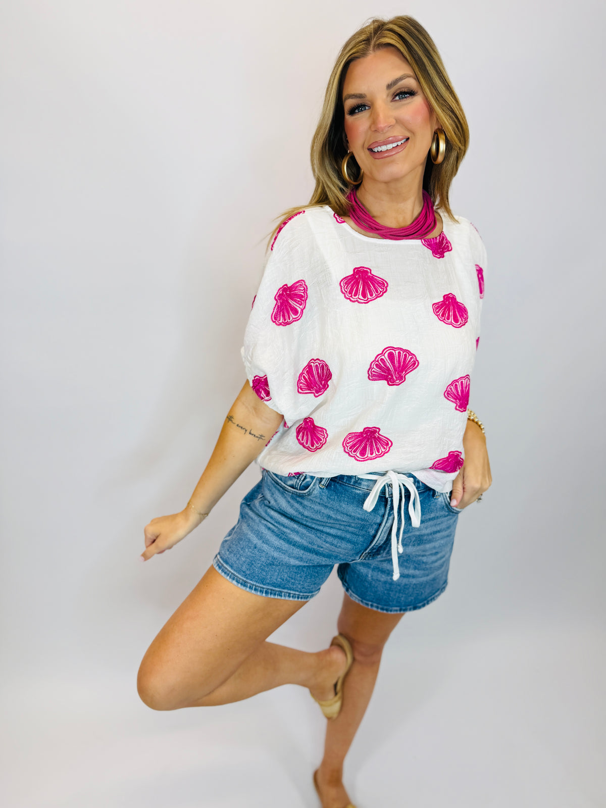 Woman wearing white seashell print top with denim drawstring shorts, smiling and posing against white background.