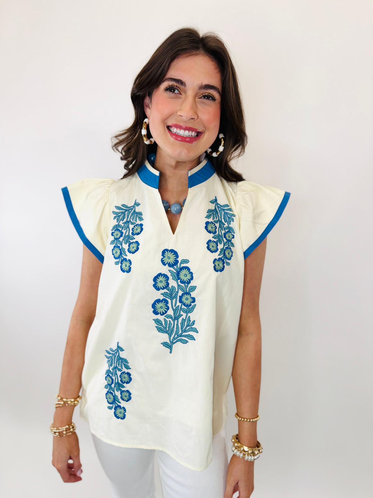 Woman wearing cream embroidered top with blue floral designs, blue collar trim, and cap sleeves