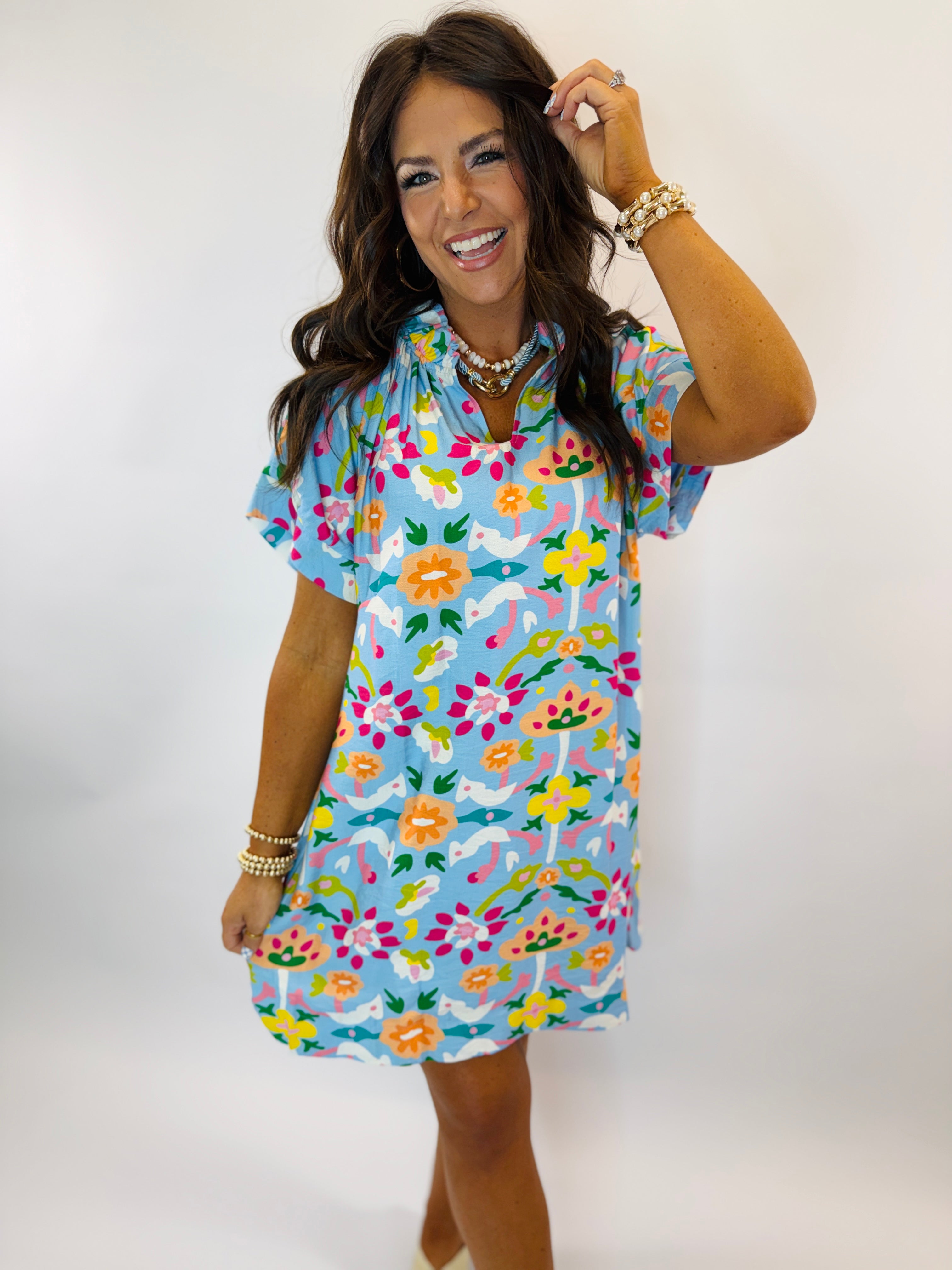 Woman wearing colorful floral print Kori dress with short sleeves and cheerful multicolored pattern on light blue background.