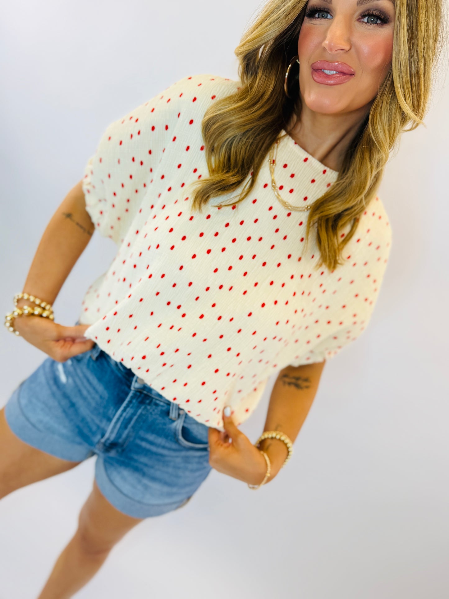 Woman wearing cream colored short-sleeve knit top with red polka dots and denim shorts
