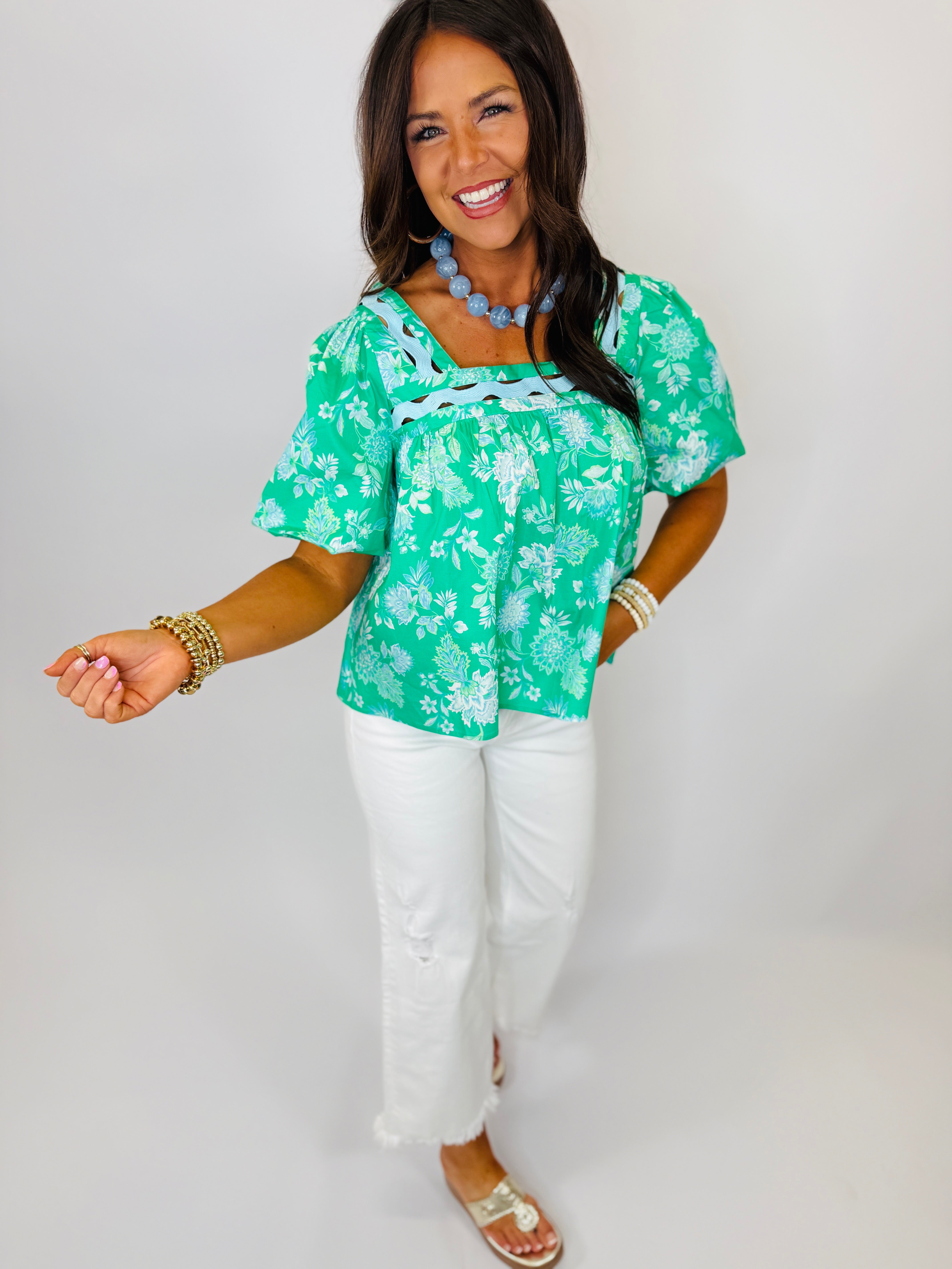 Woman wearing green floral Sydney top with white pants, blue beaded necklace, and gold accessories, smiling at camera.