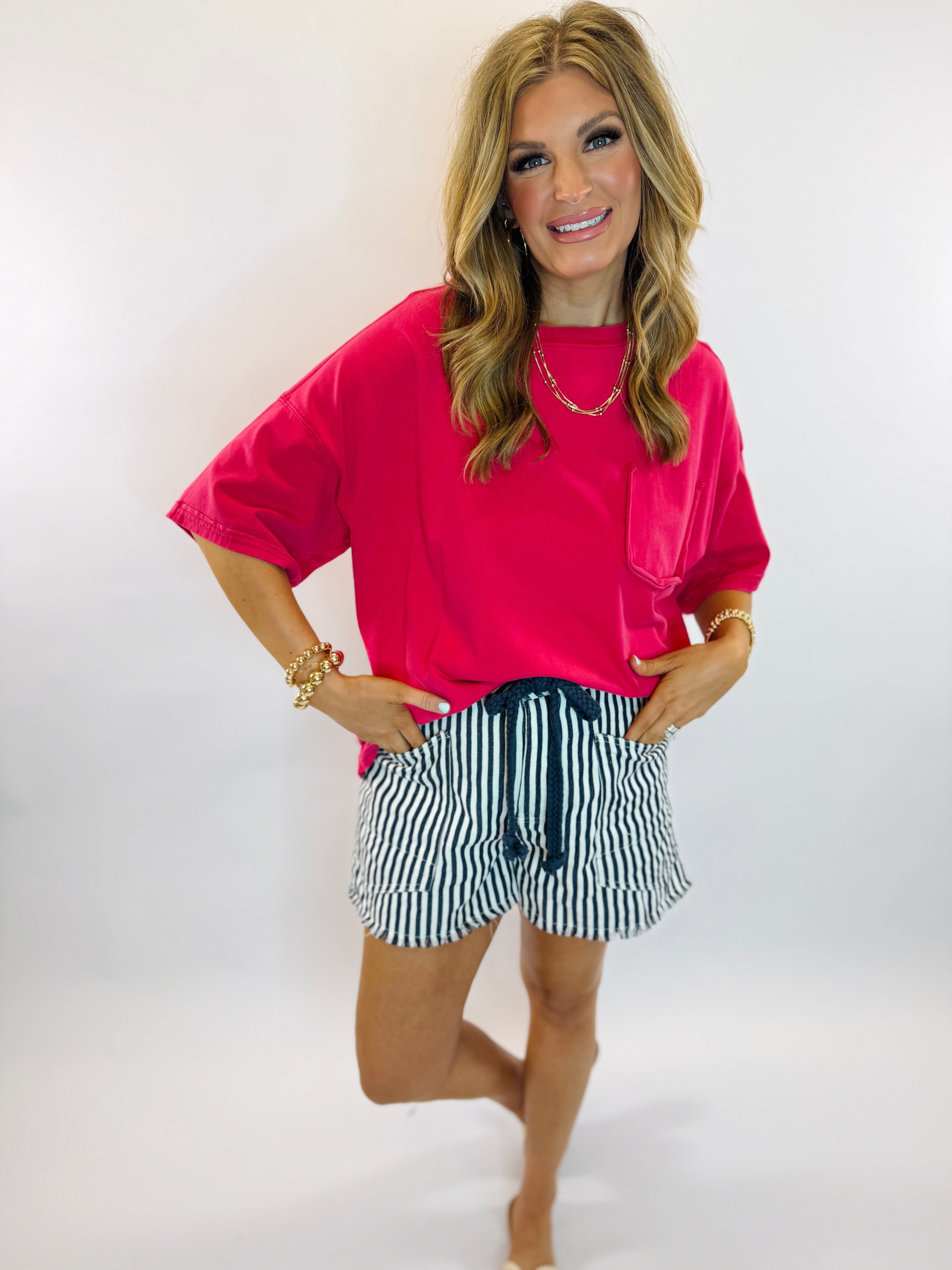 Woman wearing bright pink oversized t-shirt and navy blue striped drawstring shorts with gold jewelry accessories.