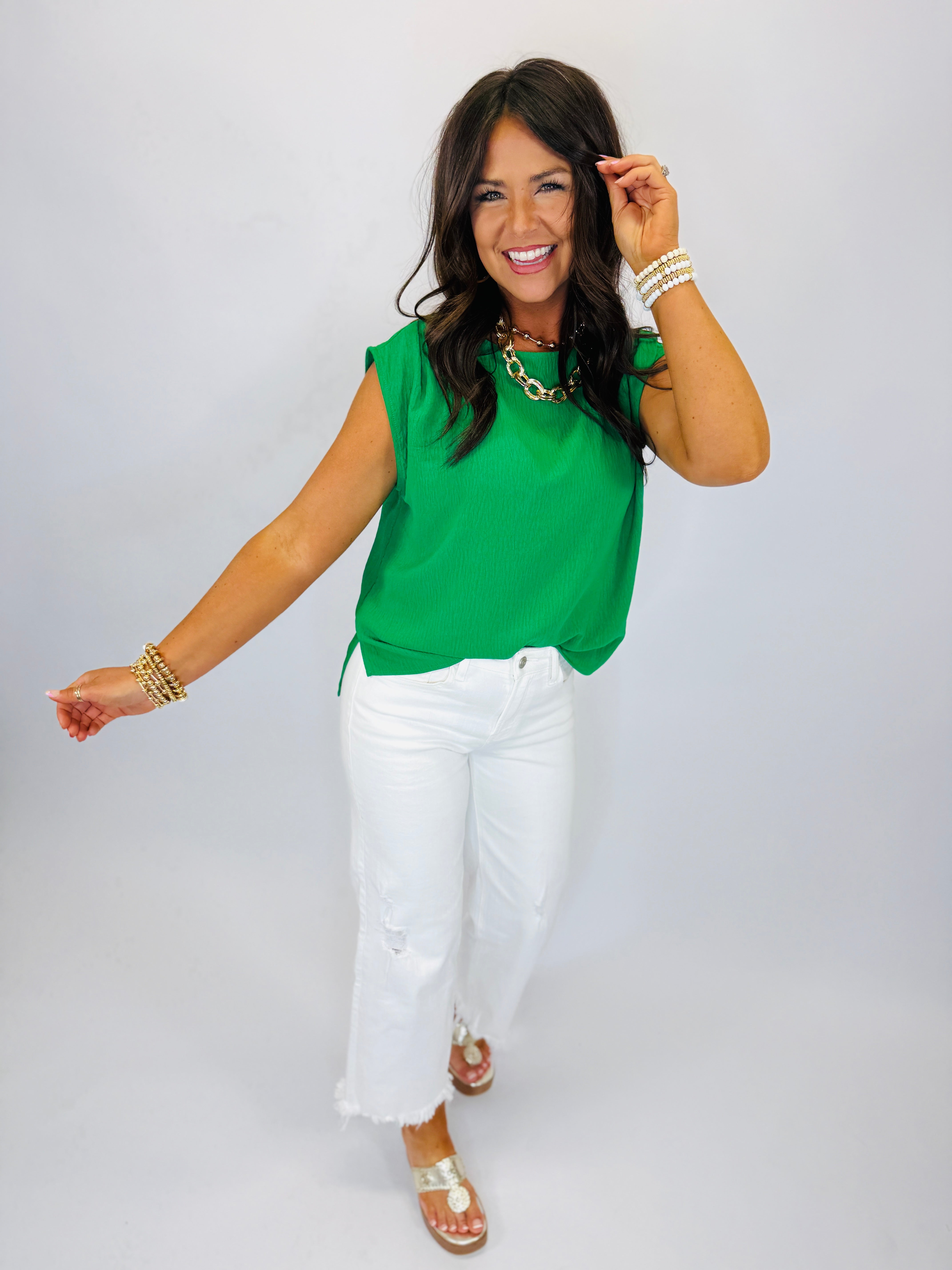 Woman wearing green sleeveless top and white wide leg jeans with gold jewelry and sandals