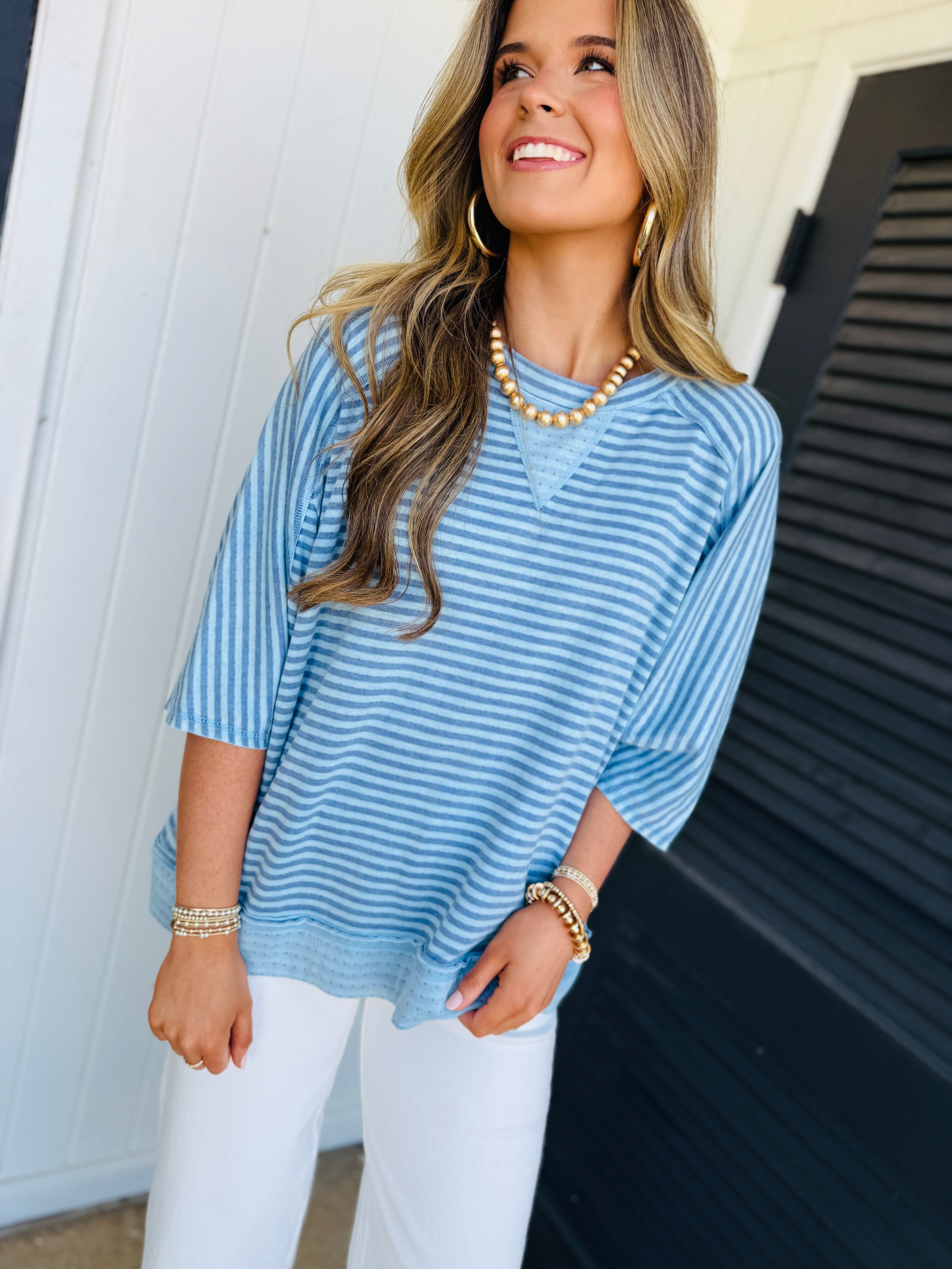 Woman wearing blue and white striped oversized pullover top with white pants and gold jewelry accessories.