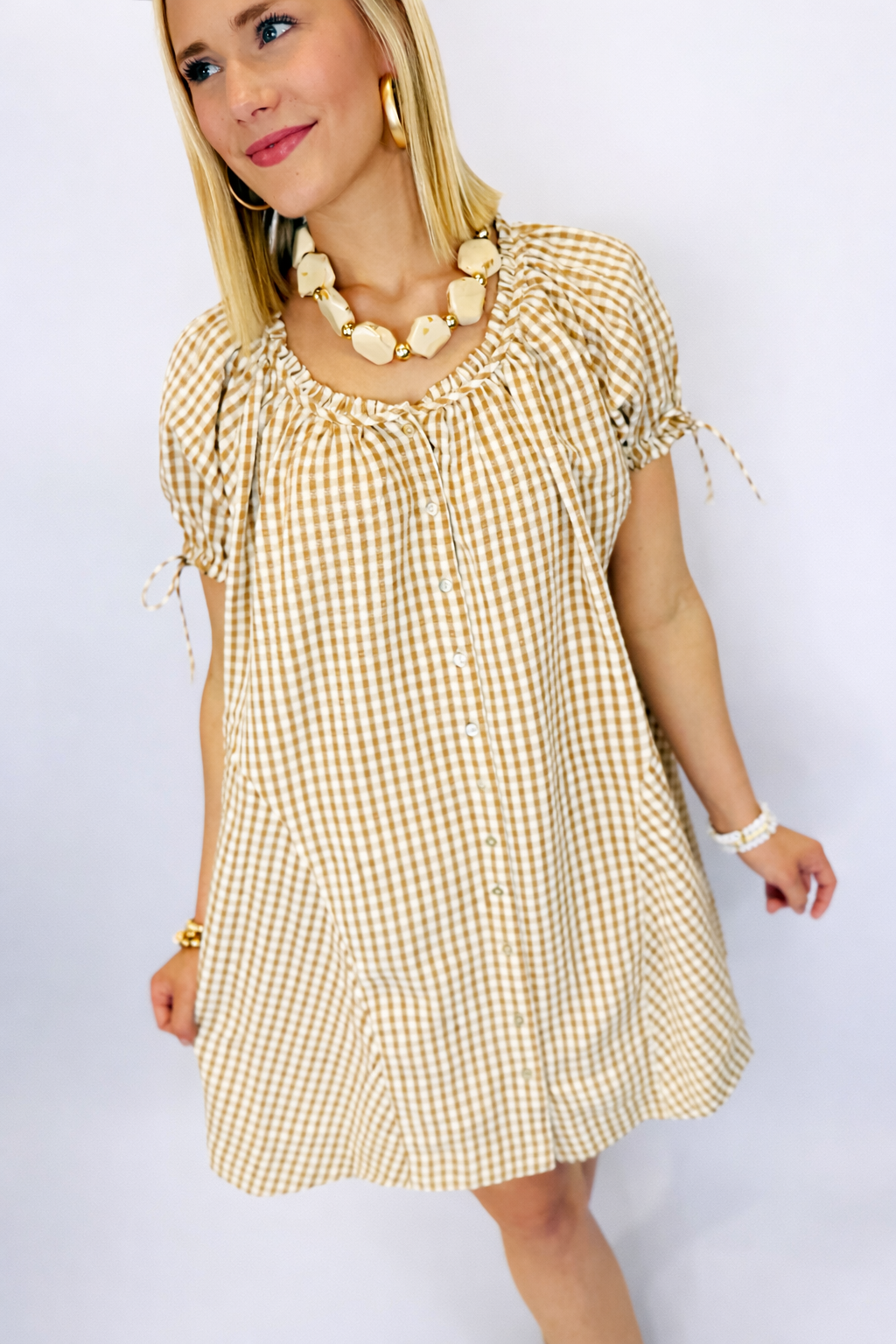 Woman wearing tan and white gingham button-front dress with puffed sleeves and bow tie details