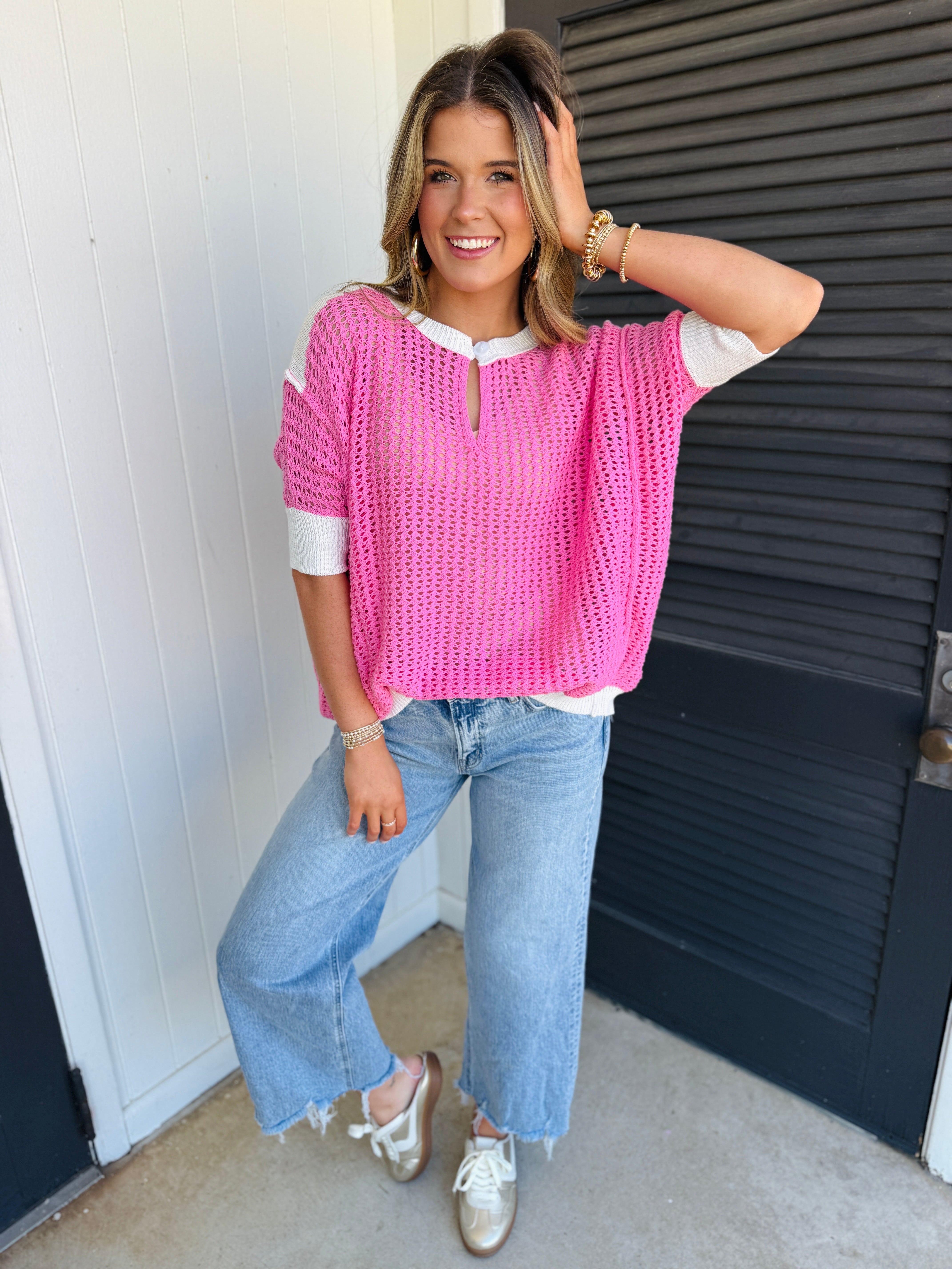Woman wearing pink knit sweater with white trim and blue jeans, smiling while posing casually.