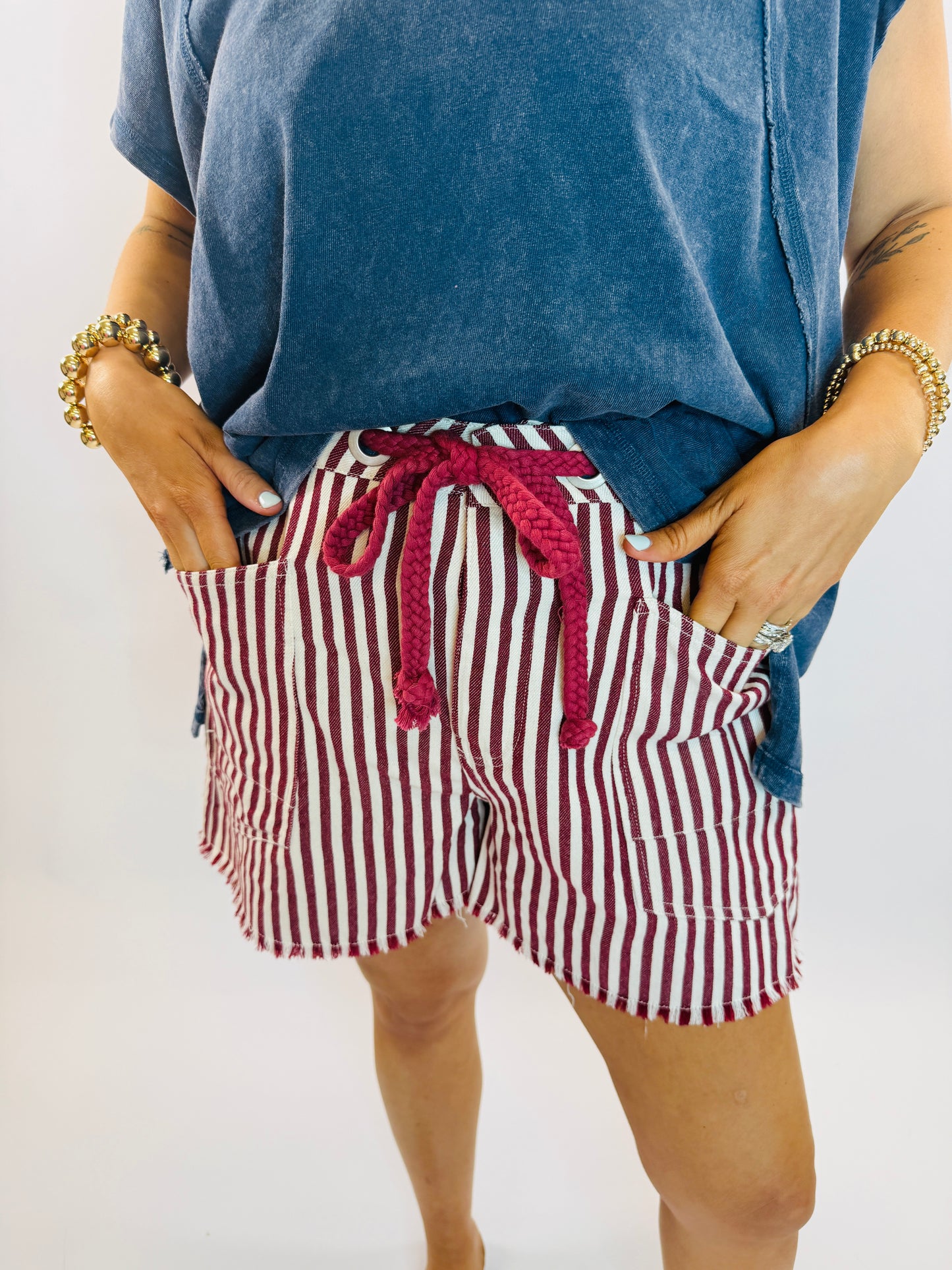 Red and white striped shorts with drawstring tie and frayed hem, styled with blue top and gold bracelets.