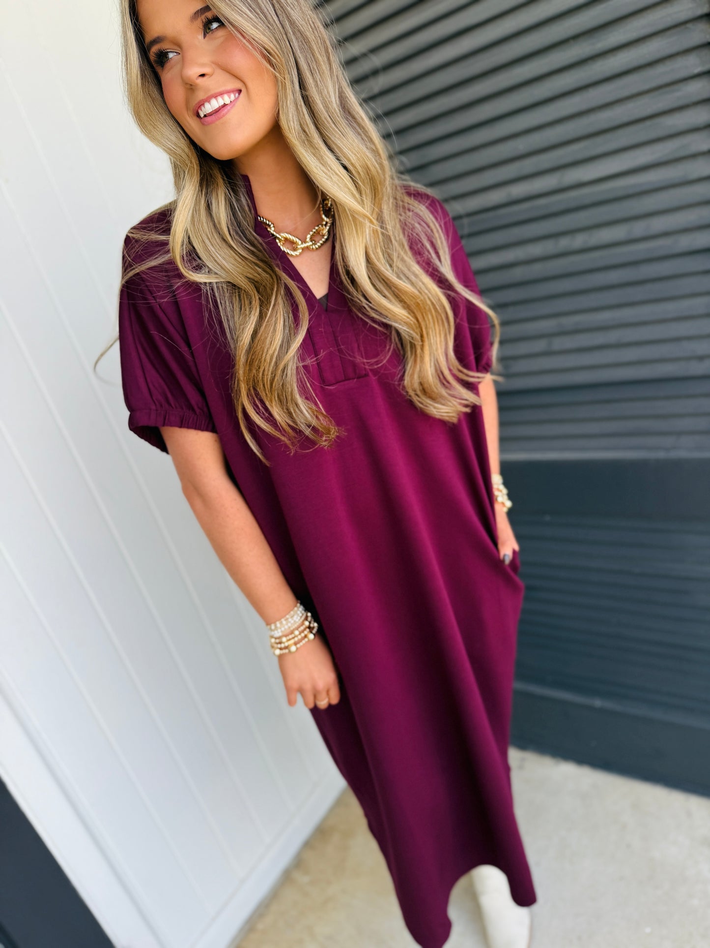 Woman wearing burgundy flowy v-neck dress with short sleeves, styled with gold chain necklace and pearl bracelet.