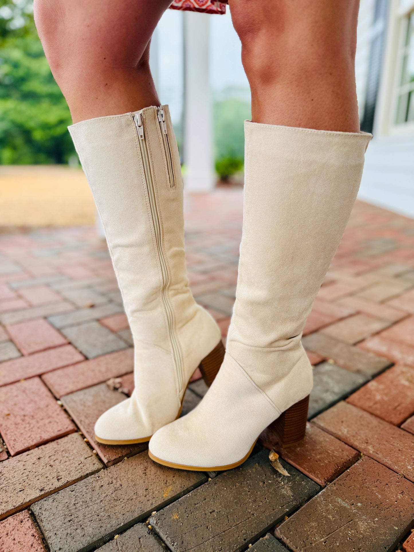 Cream-colored knee-high boots with wooden block heels and side zippers on brick patio
