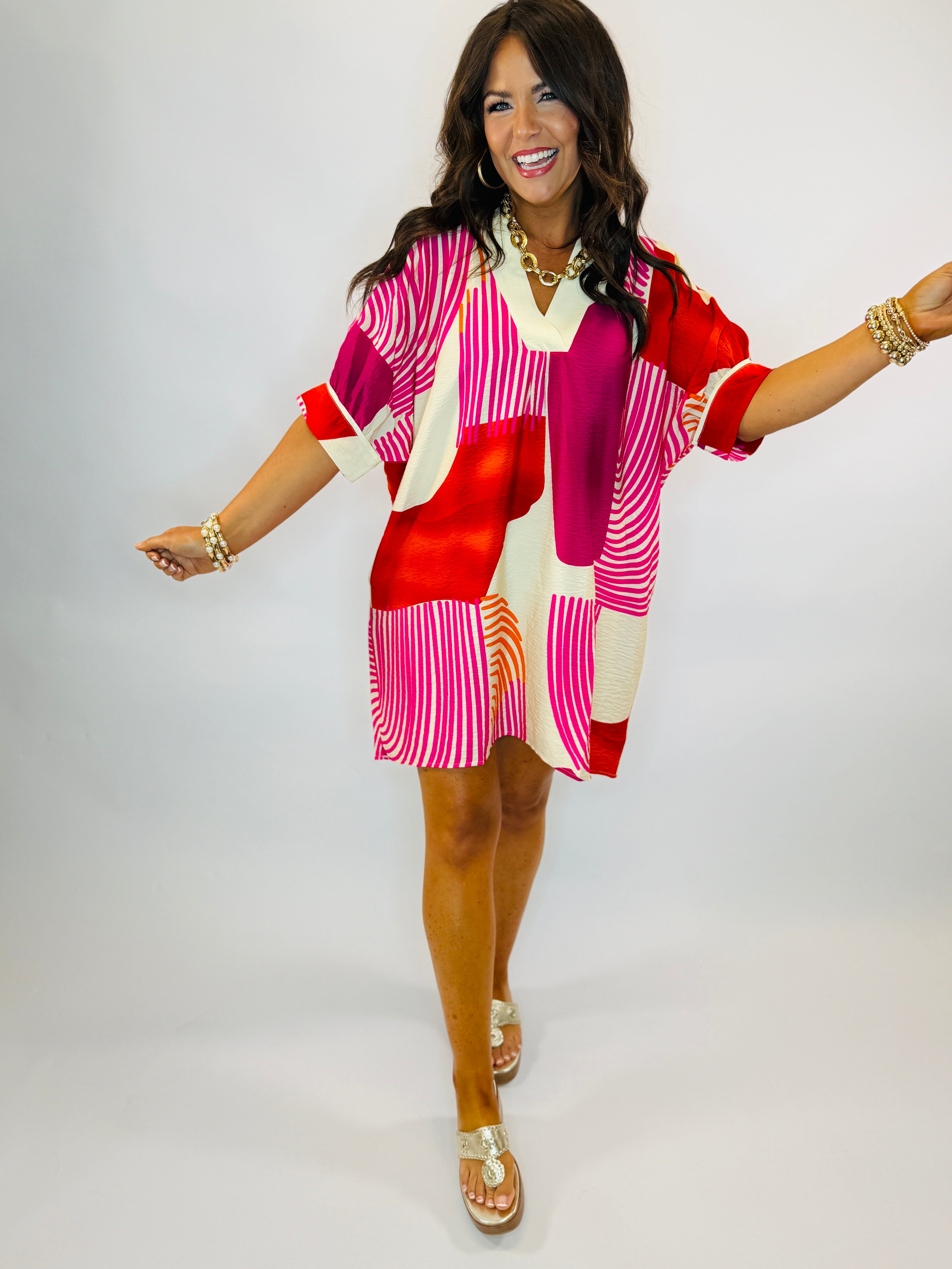 Woman wearing colorful Hannah dress with pink, red, and white geometric pattern, three-quarter sleeves, and gold jewelry a...