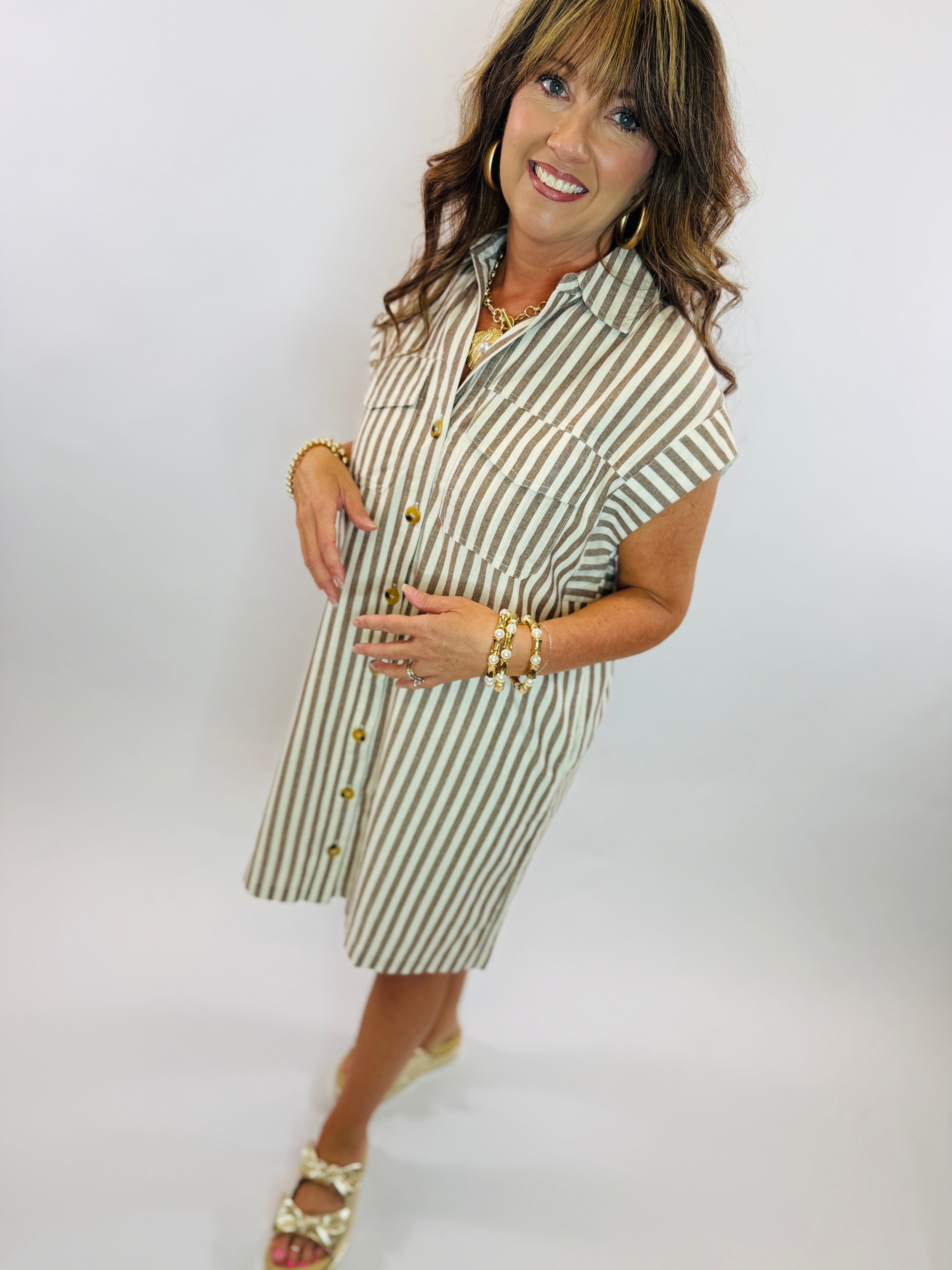 Woman wearing olive green and white striped button-front shirt dress with gold jewelry and sandals