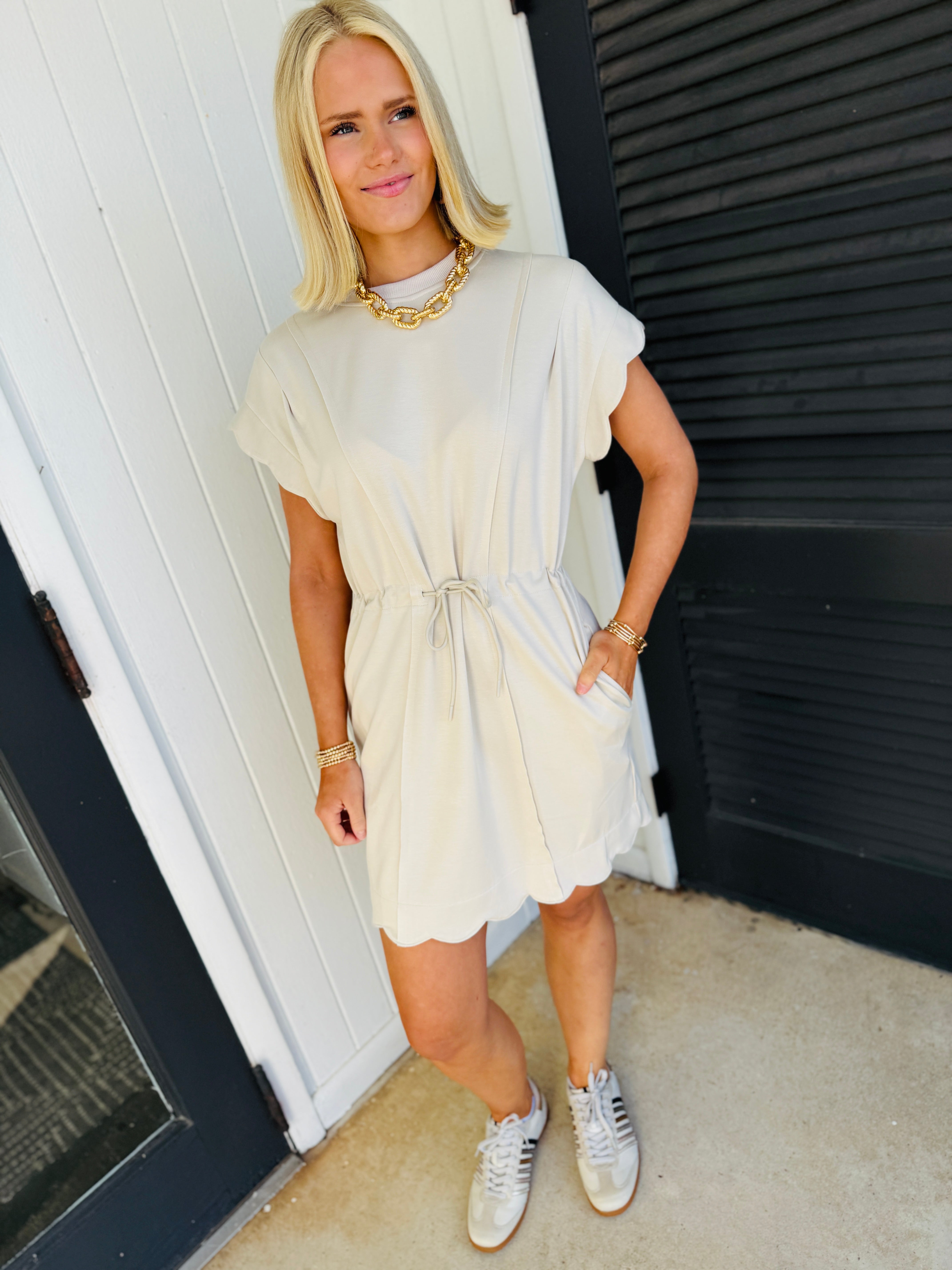 Woman wearing beige short-sleeve dress with tie waist, gold chain necklace, and white sneakers