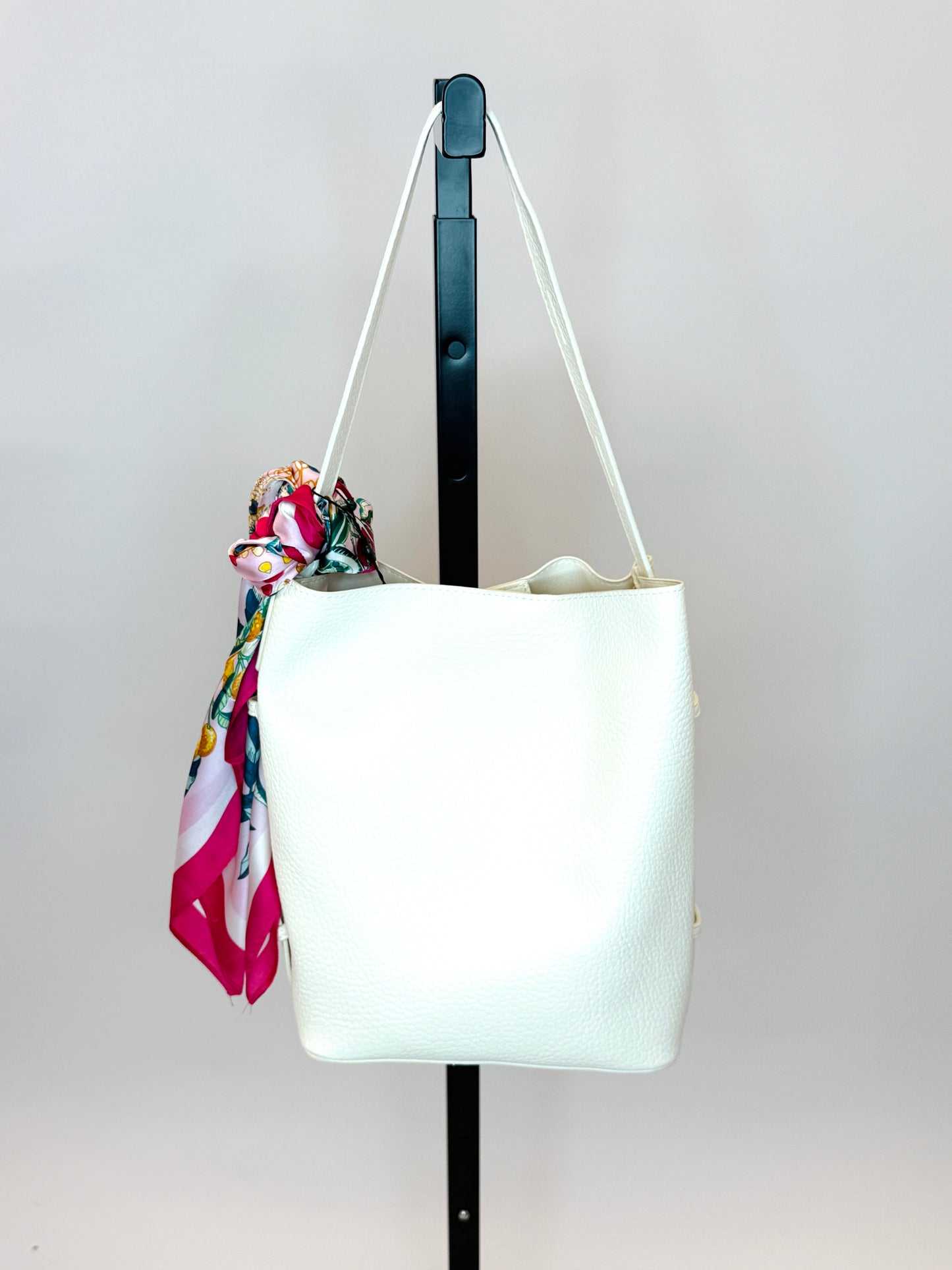White leather handbag with adjustable straps and colorful floral scarf accessory hanging on display stand.