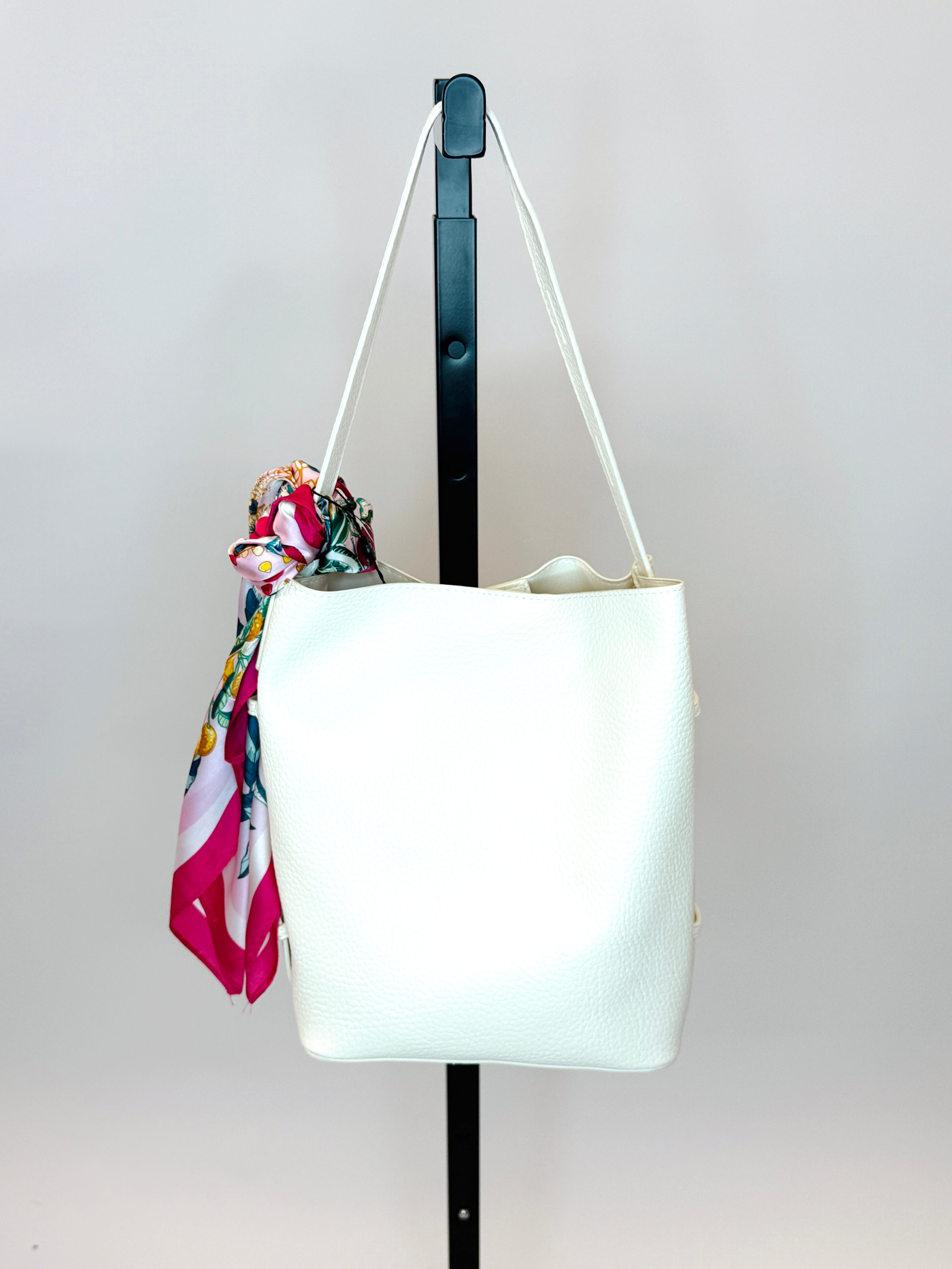 White leather handbag with adjustable straps and colorful floral scarf accessory hanging on display stand.