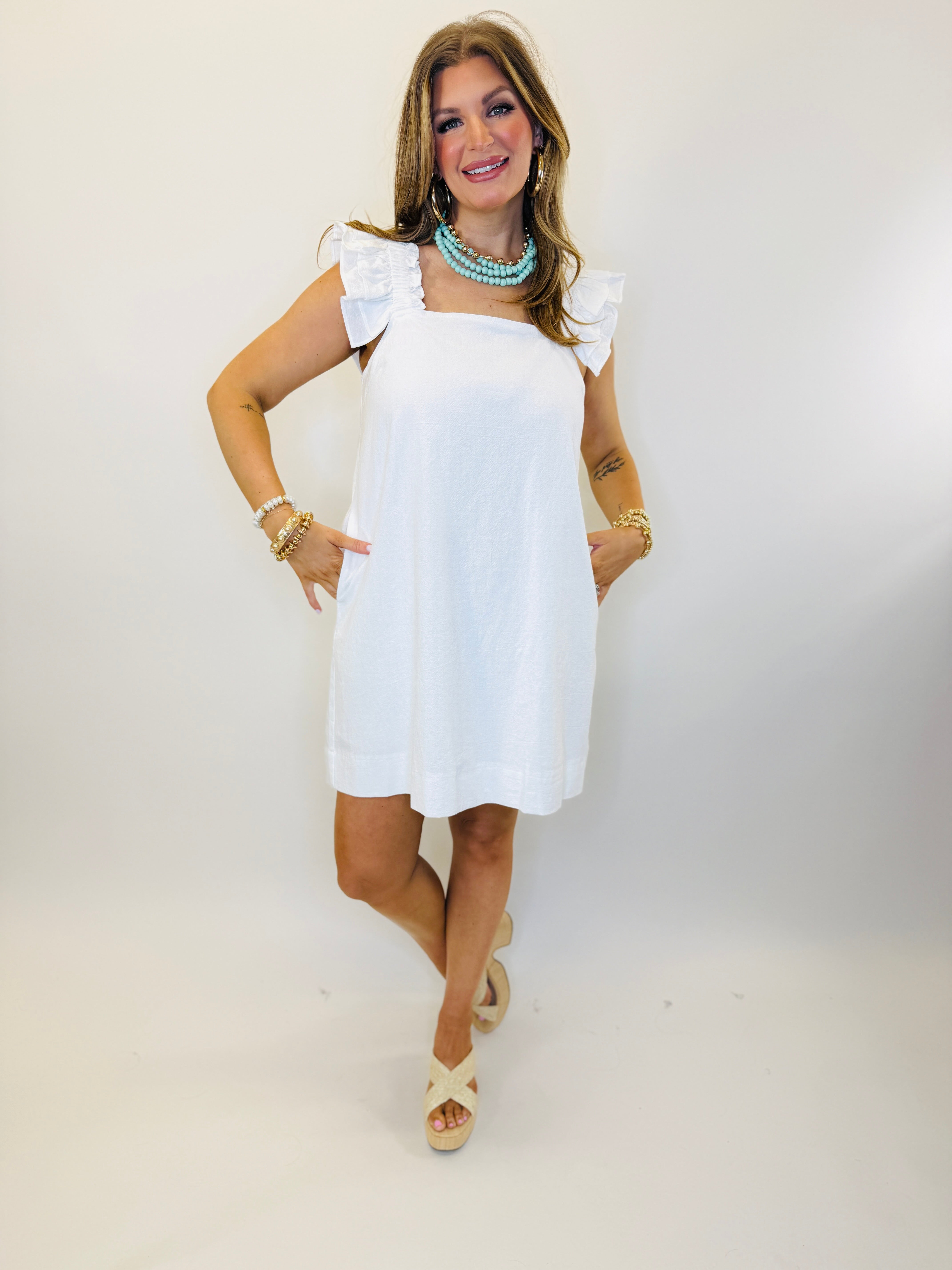 White sleeveless dress with ruffle shoulder straps, knee-length, worn with turquoise necklace and gold bracelets.