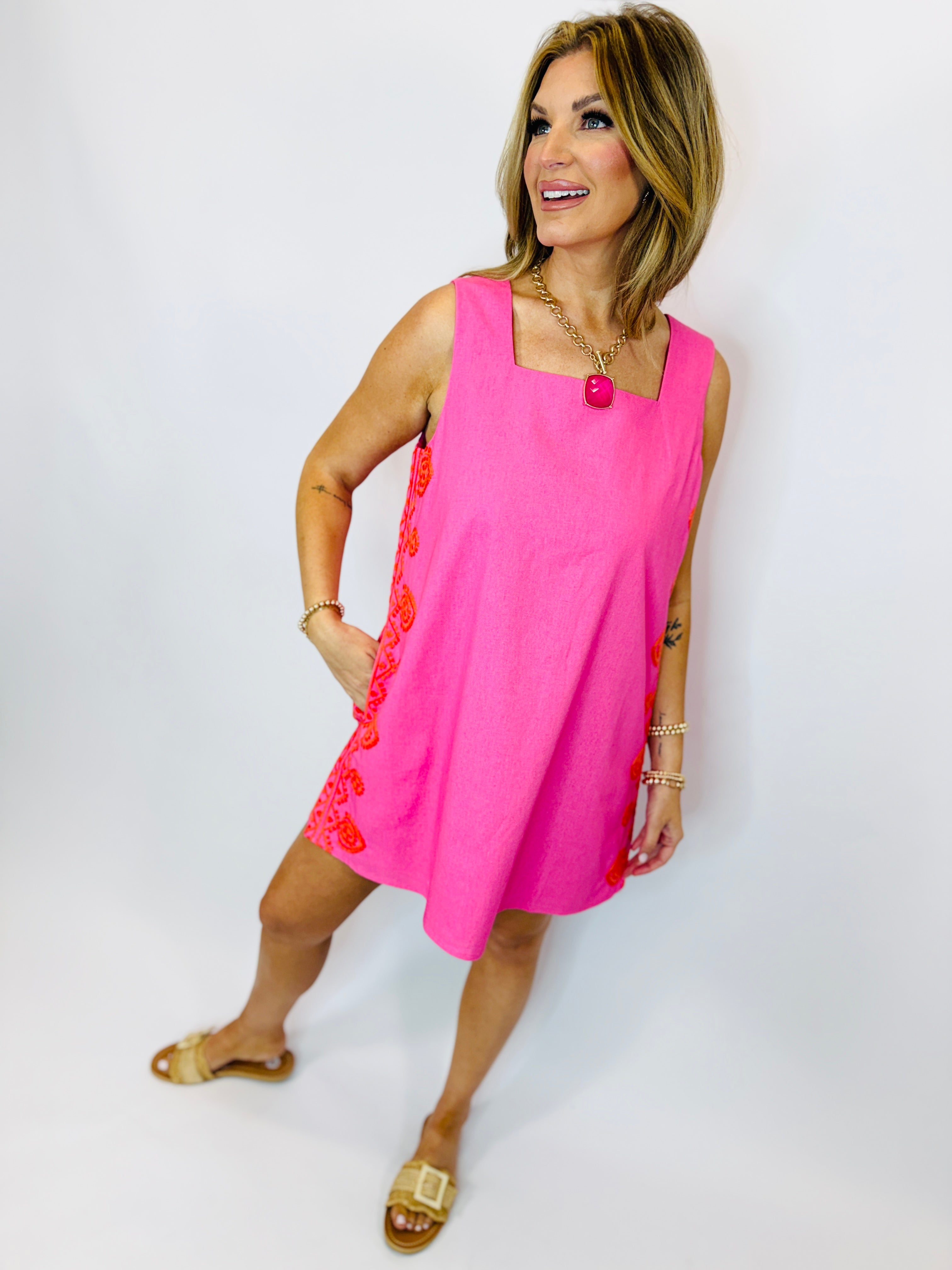 Woman wearing bright pink sleeveless Marlene dress with red floral embroidery detail and gold sandals.