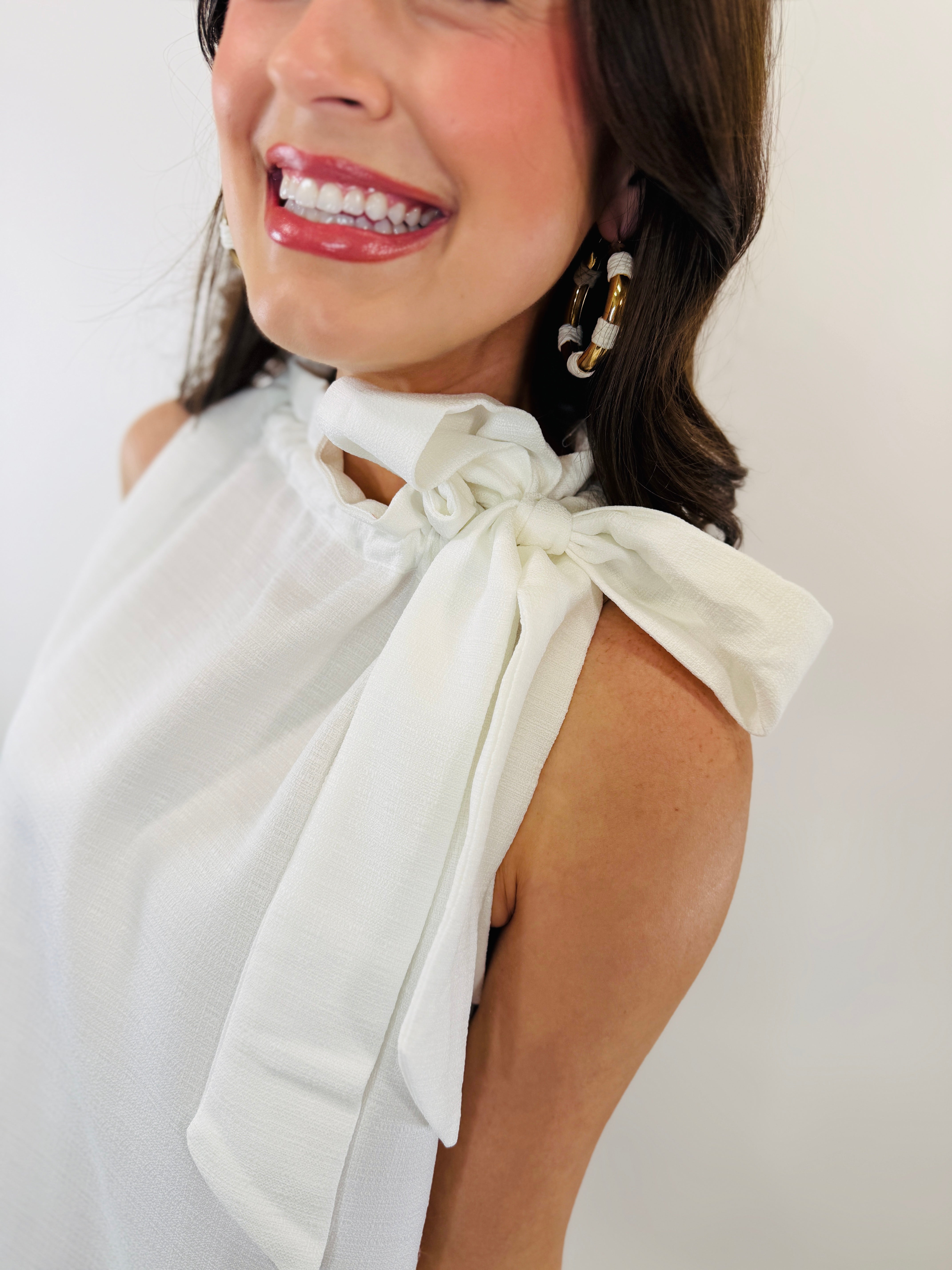 White sleeveless dress with decorative bow tie detail at shoulder, worn by smiling woman with statement earrings.
