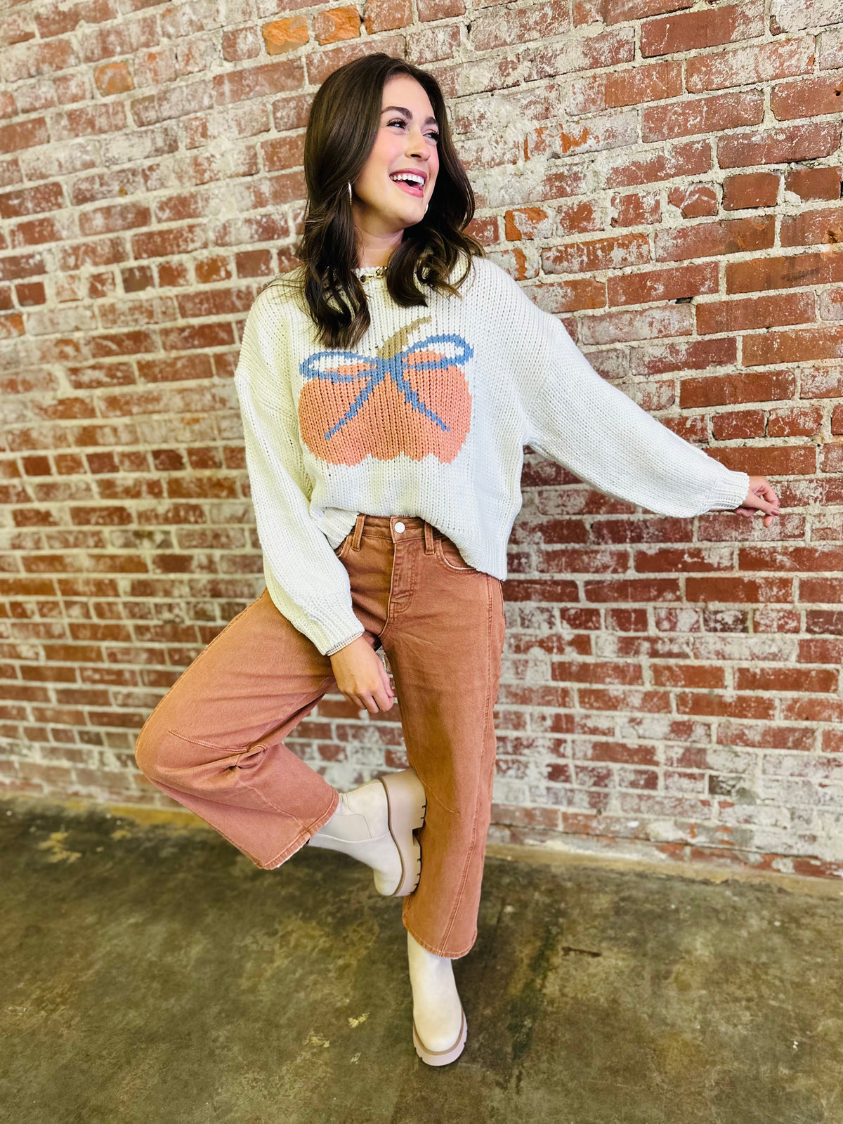 Woman wearing cream knit sweater with orange pumpkin design and blue bow, paired with brown jeans.